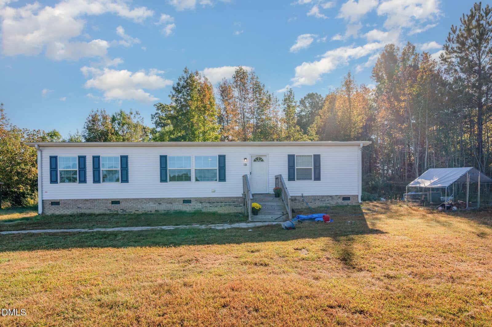 A single-story white manufactured home with a brick foundation sits on a grassy lot. There are steps leading to the front door, toys on the lawn, and trees in the background under a partly cloudy sky.