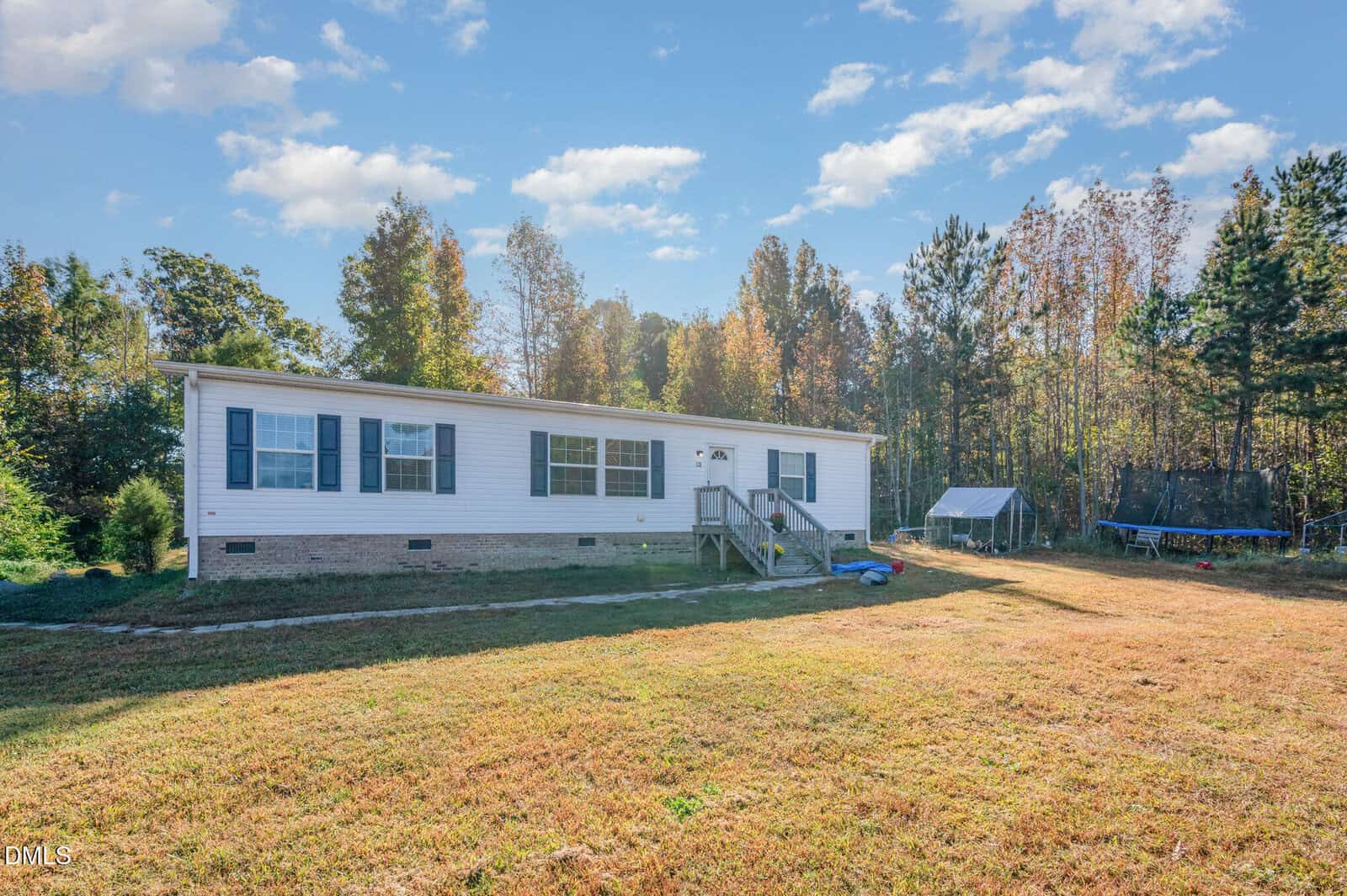 A single-story white manufactured home with a small front porch sits on a grassy yard bordered by trees. A trampoline and a small structure are visible in the background. The sky is partly cloudy.