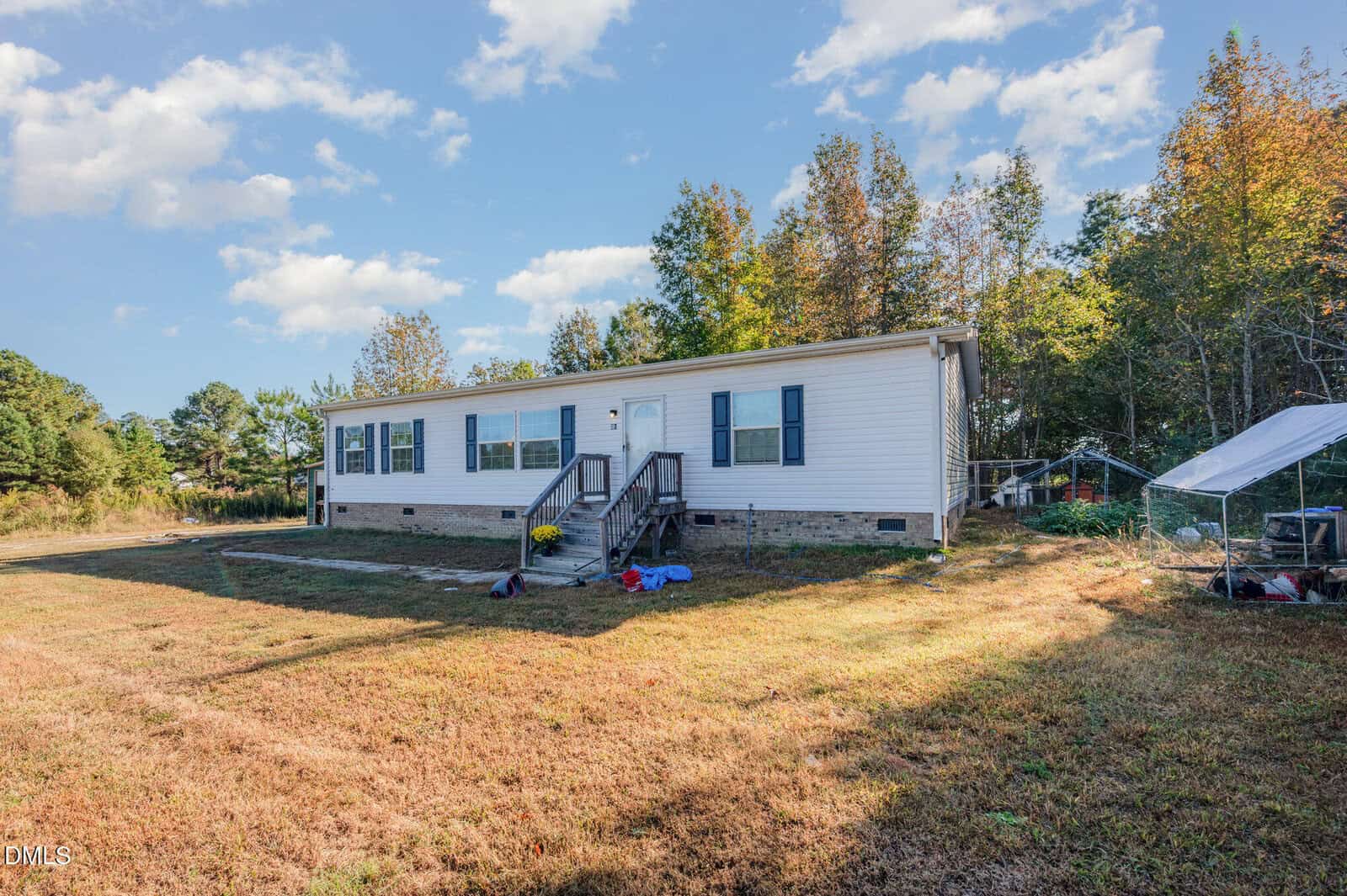 A single-story manufactured home with white siding and blue shutters sits on a large grassy lot. There are trees in the background, and some outdoor items and a small greenhouse are visible near the house.