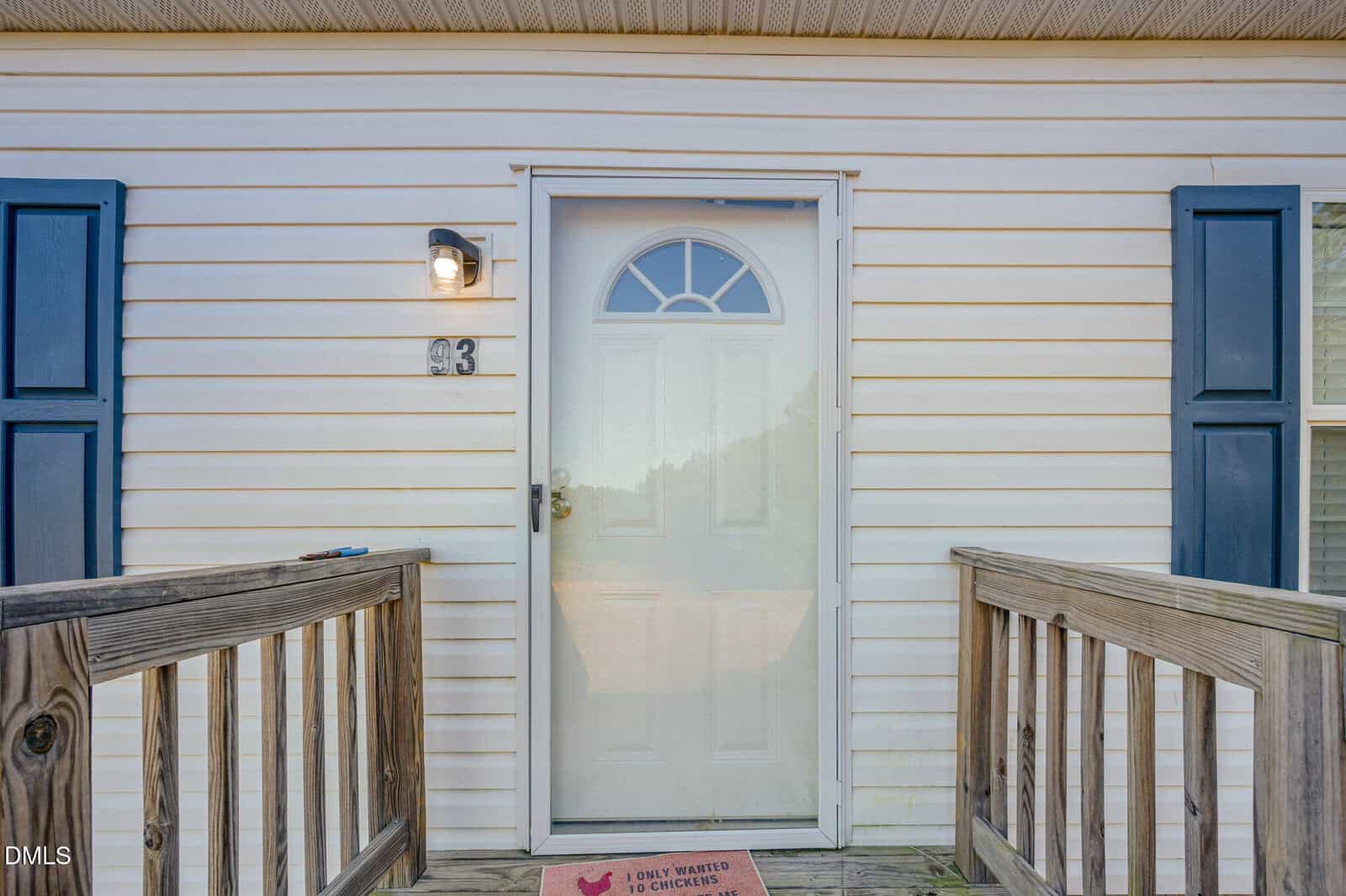 A white front door with a semicircular window at the top, flanked by blue shutters, with a porch light, house number 43, and a doormat. Wooden railings lead up to the entrance.