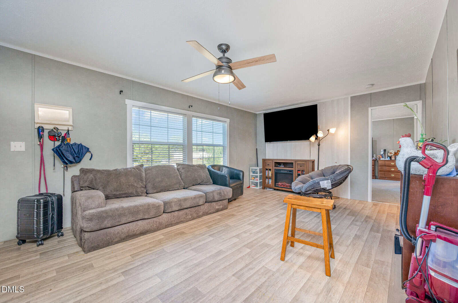 A living room with a gray couch, wooden chair, TV above a fireplace, ceiling fan, and large window. Cleaning tools and bags are against one wall. An open doorway leads to another room with a dresser.
