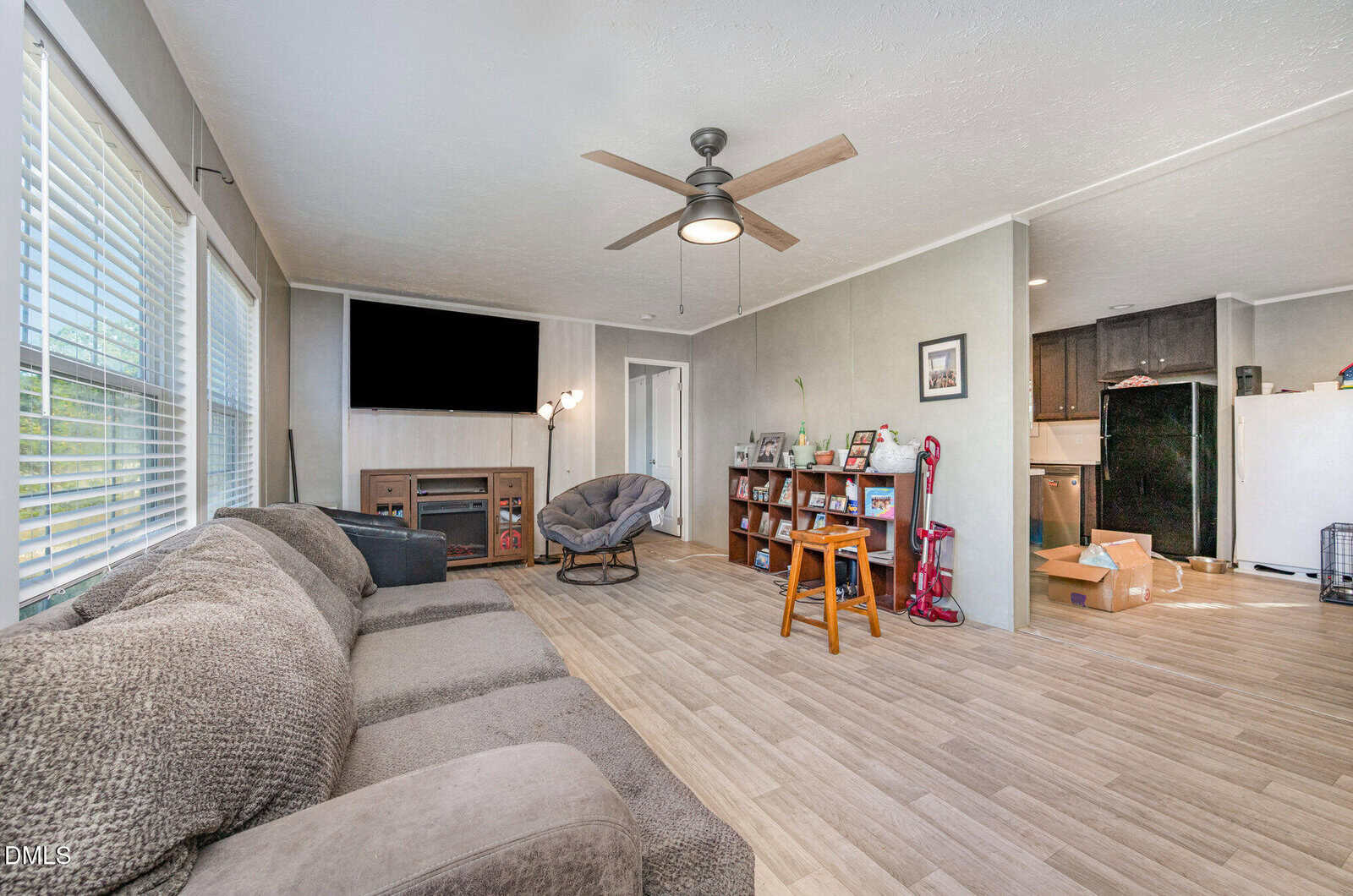 A living room with a gray sectional sofa, a ceiling fan, a TV on a fireplace, a round chair, a wooden bookshelf, a vacuum, and an open view into a kitchen with black appliances and unpacked boxes.