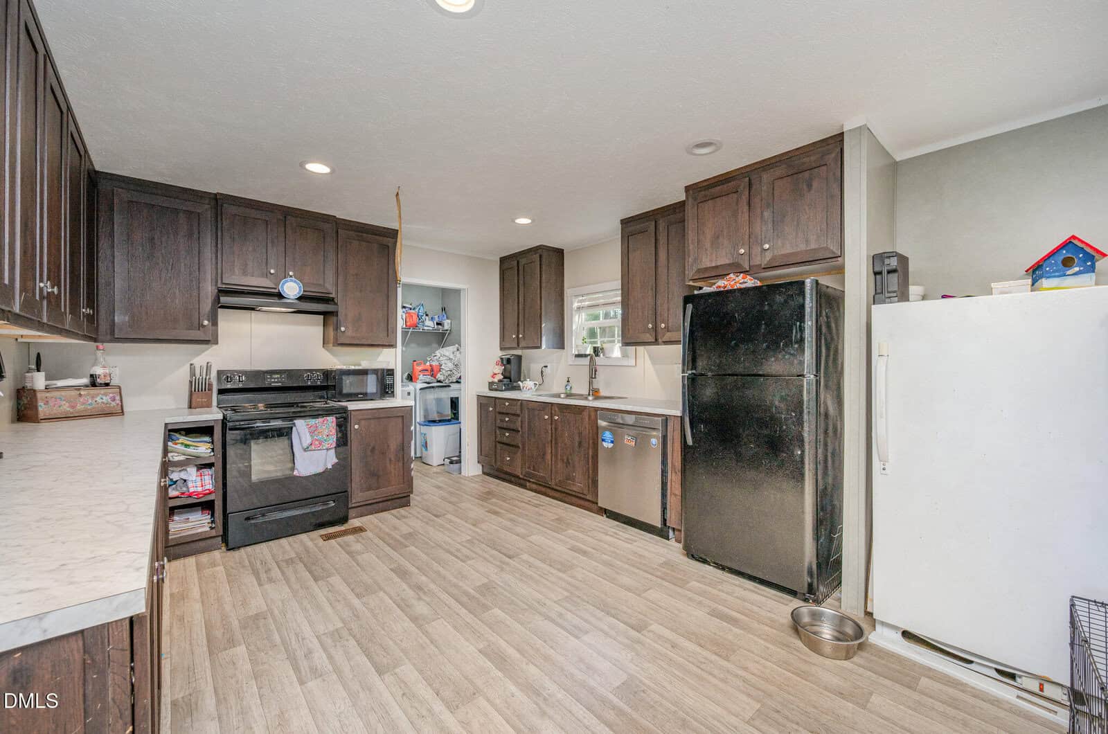 A kitchen with dark wood cabinets, black appliances, a white refrigerator, light wood flooring, a white marble countertop, and an assortment of household items on counters and open shelves. A pet bowl is on the floor.
