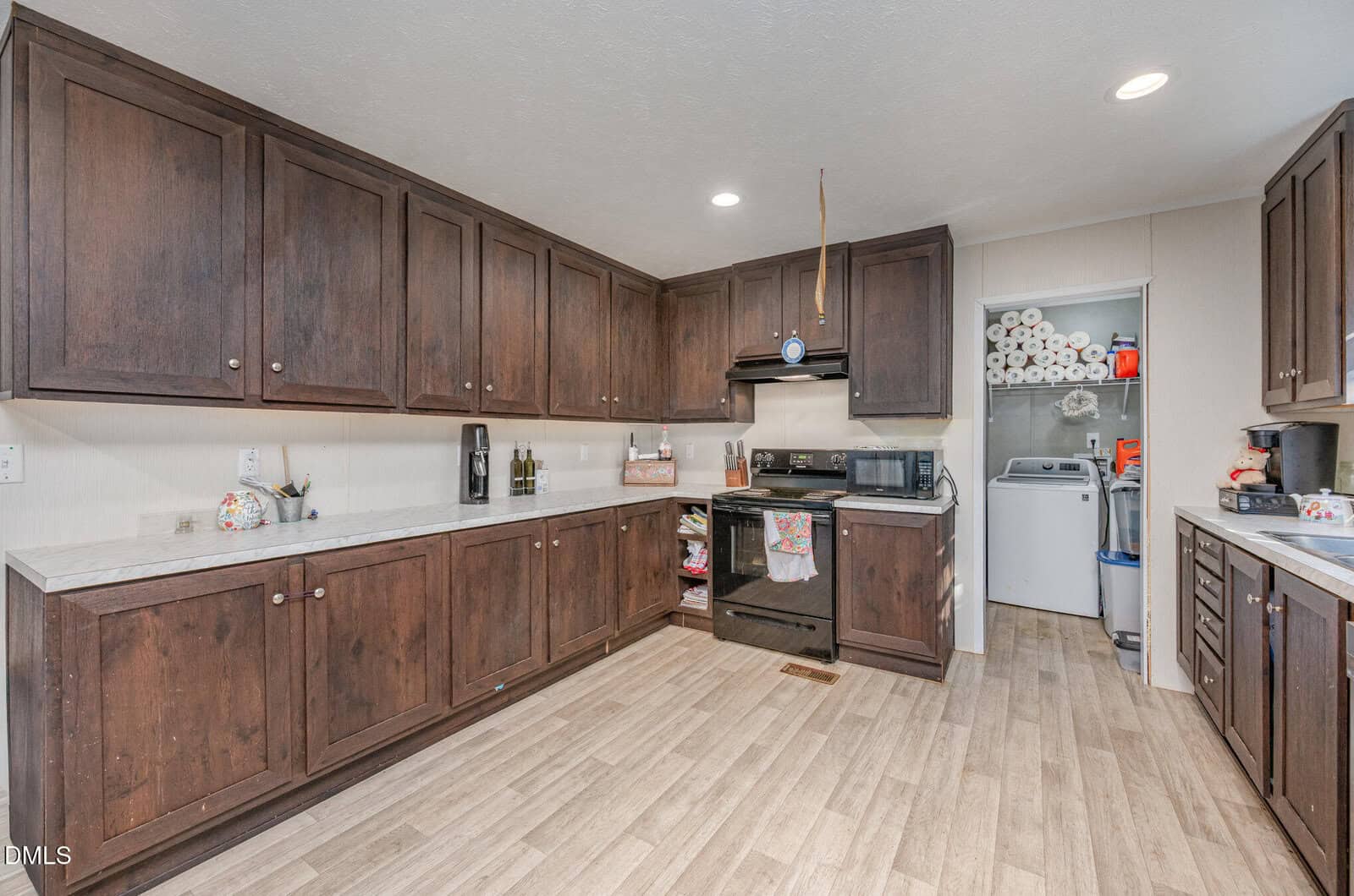 Spacious kitchen with dark wooden cabinets, light countertops, stove, microwave, and coffee maker. In the background, laundry area with washer, dryer, and stacked paper towels is visible through an open doorway.