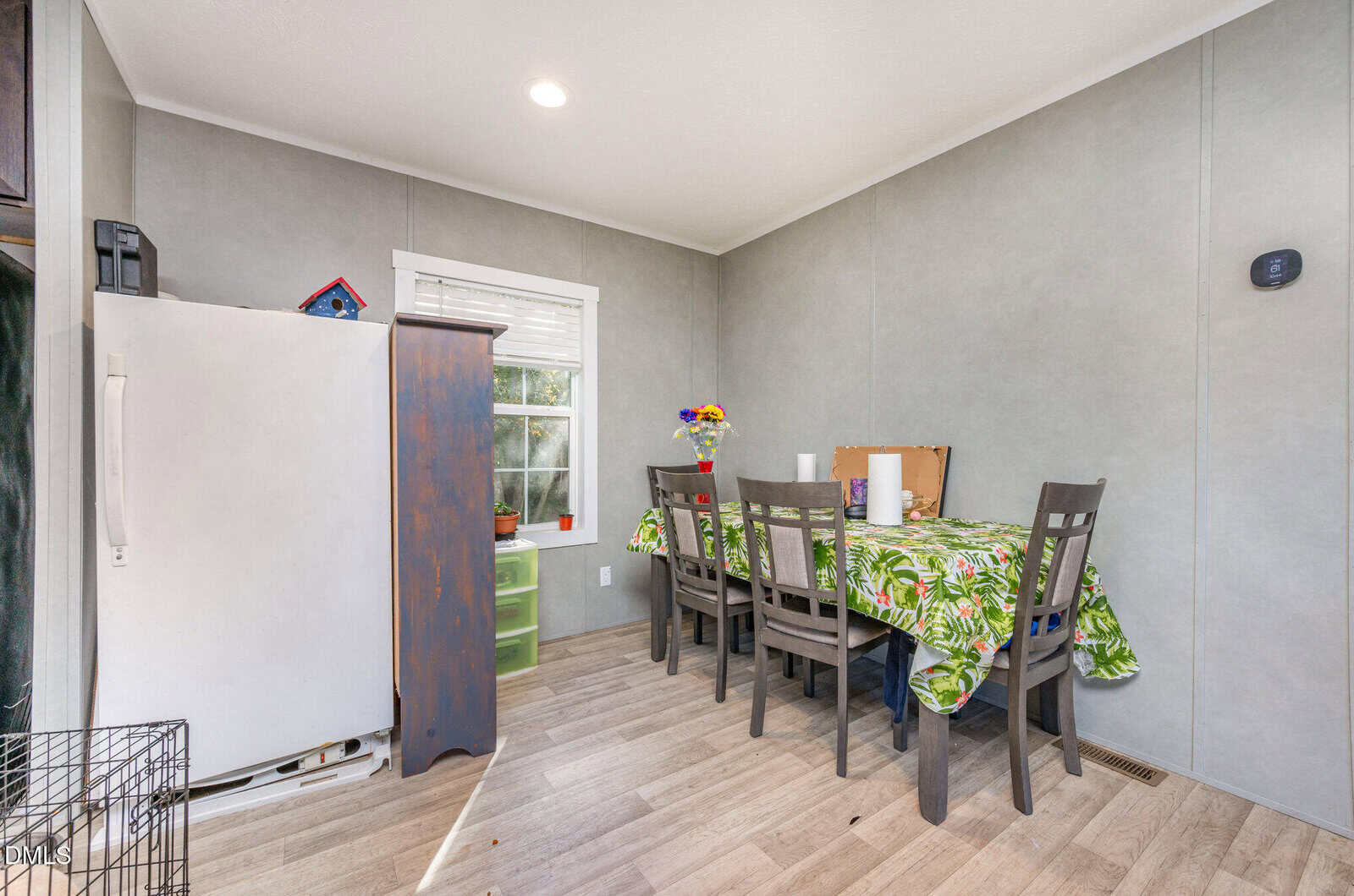 Small dining area with a table covered by a green leafy tablecloth, four chairs, a white refrigerator, a tall blue cabinet, and a window letting in natural light. The floor is light wood, and walls are gray.