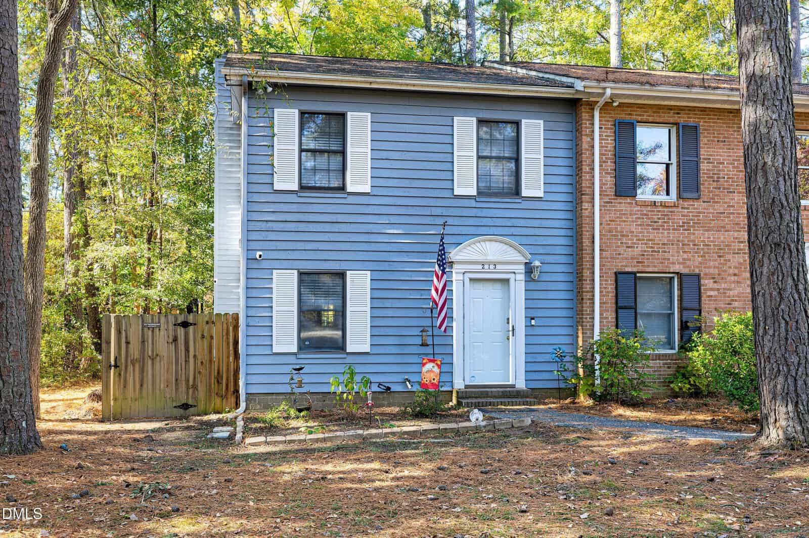 A two-story blue townhouse with white shutters and a white door stands proudly at 213 12th Street in Butner, featuring an American flag and a small garden in front. A wooden fence is on the left, with trees and pine needles covering the yard.