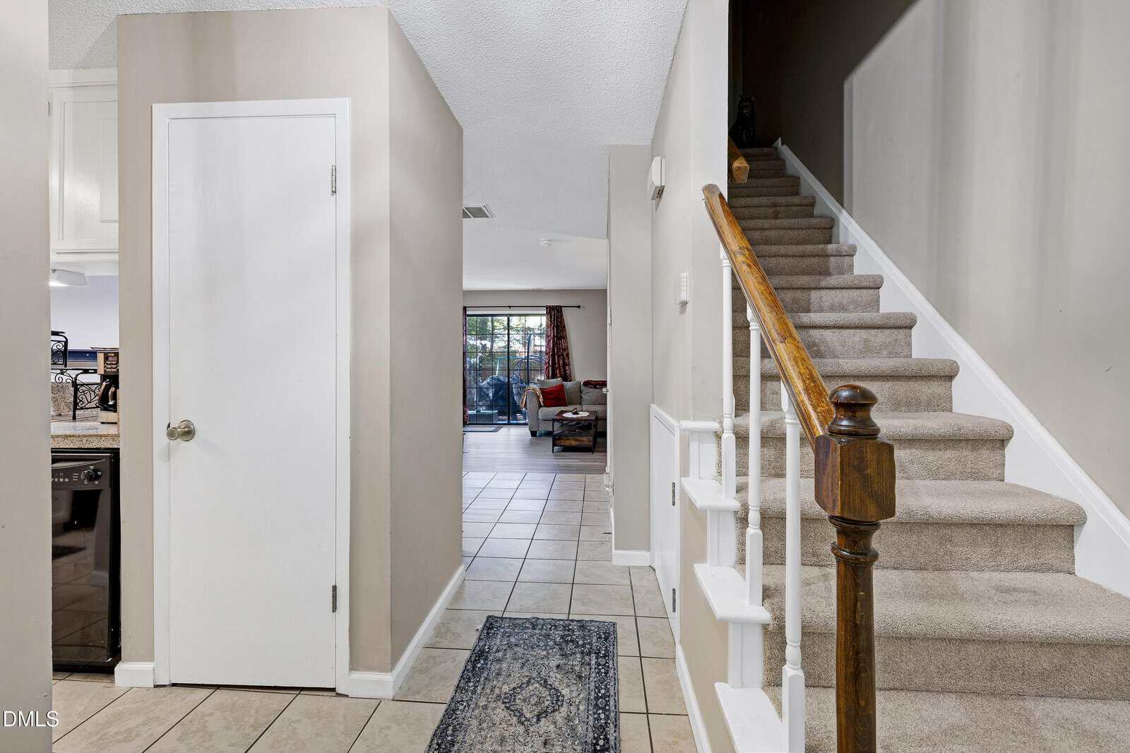 A carpeted staircase with a wooden handrail is on the right. To the left, there is a closed white door and a tiled hallway leading to a living area with a sliding glass door and visible furniture.