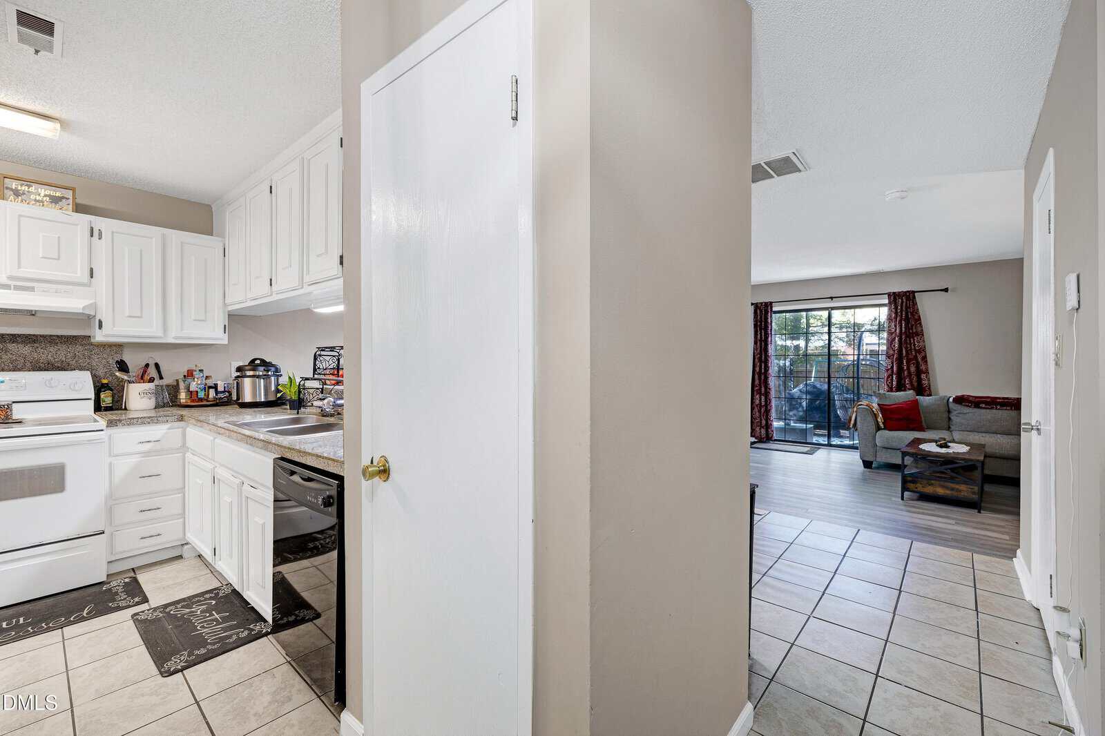 A view of a kitchen with white cabinets and appliances to the left, and a living room with a couch and glass doors leading outside to the right, separated by a small hallway with beige walls and tile floors.