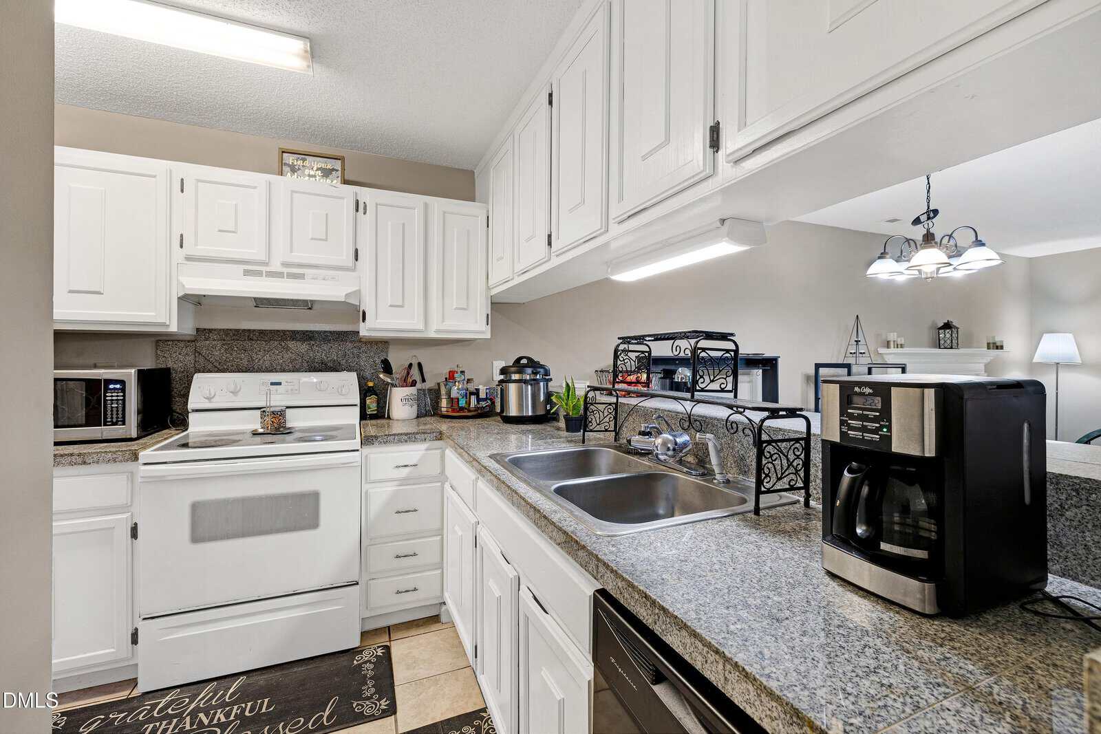 A kitchen with white cabinets, granite countertops, a white stove and oven, microwave, stainless steel sink, coffee maker, and various small appliances. Decorative items and utensils are placed on the counters and shelves.