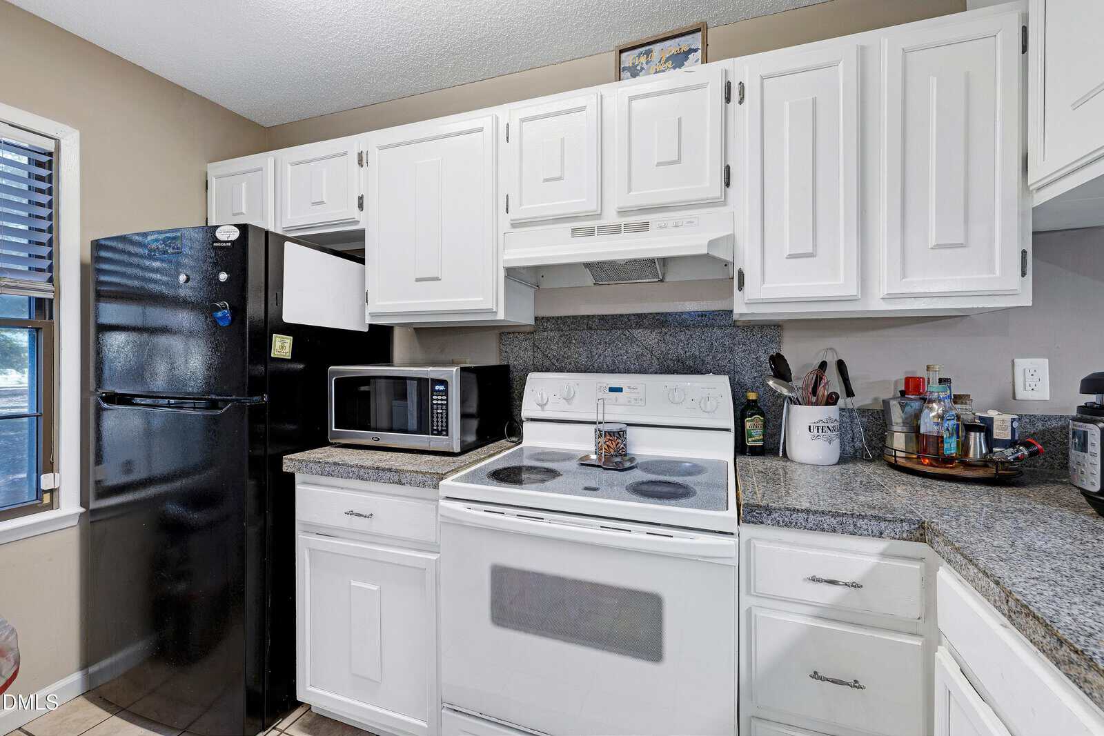 A kitchen with white cabinets, a black refrigerator, a white electric stove with oven, a microwave, granite countertops, and various kitchen items on the counters. A window is visible to the left.