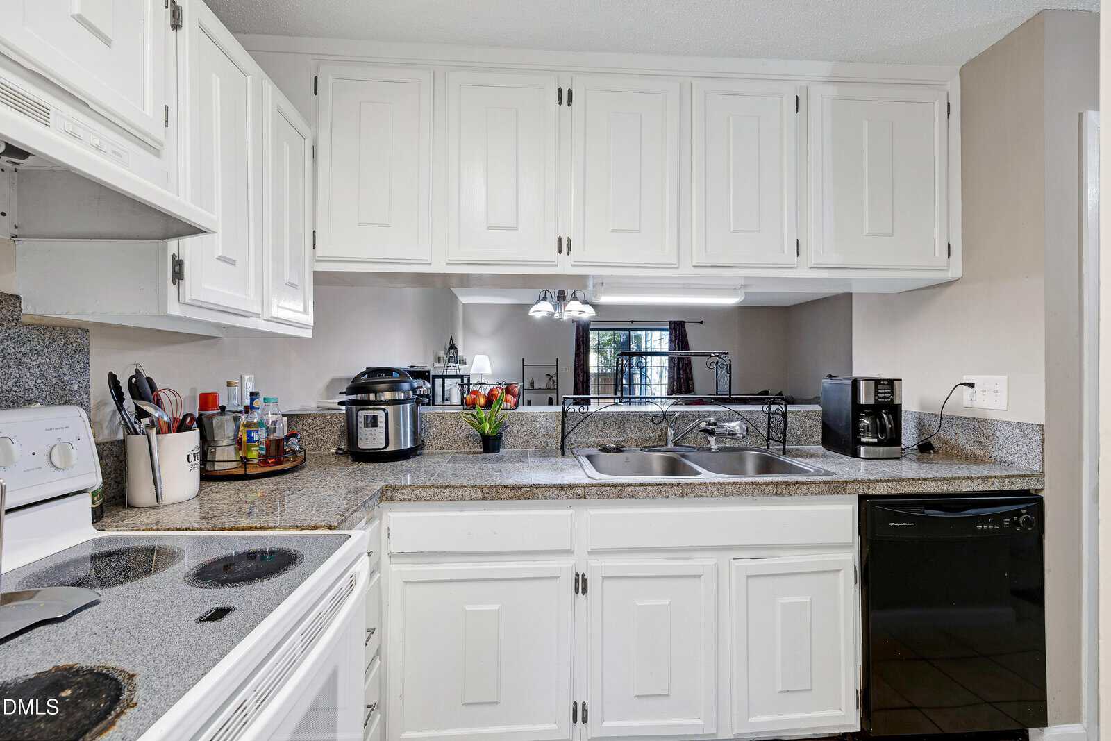 A kitchen with white cabinets, granite countertops, and stainless steel double sink. Appliances include a stove, microwave, slow cooker, toaster, and dishwasher. Small plants and kitchen items are on the counter.