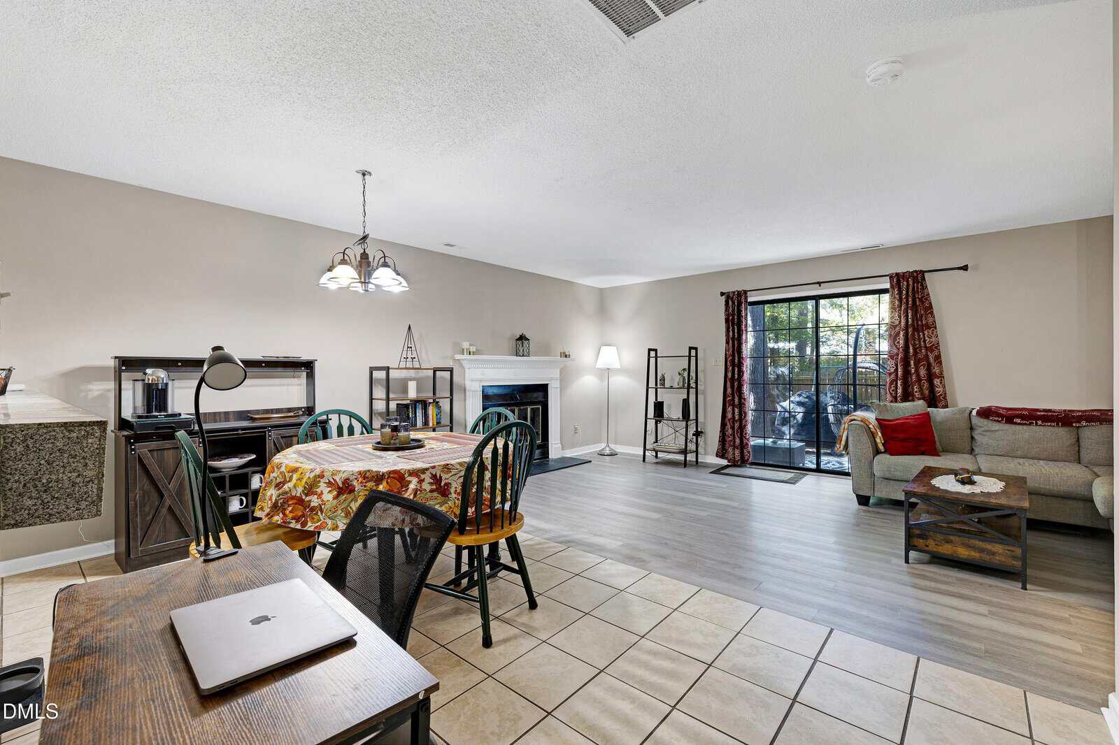 Open-plan living and dining area with tiled and wood floors, a dining table with a floral tablecloth, desk with laptop, couch, coffee table, shelves, fireplace, and sliding glass door with curtains leading outside.