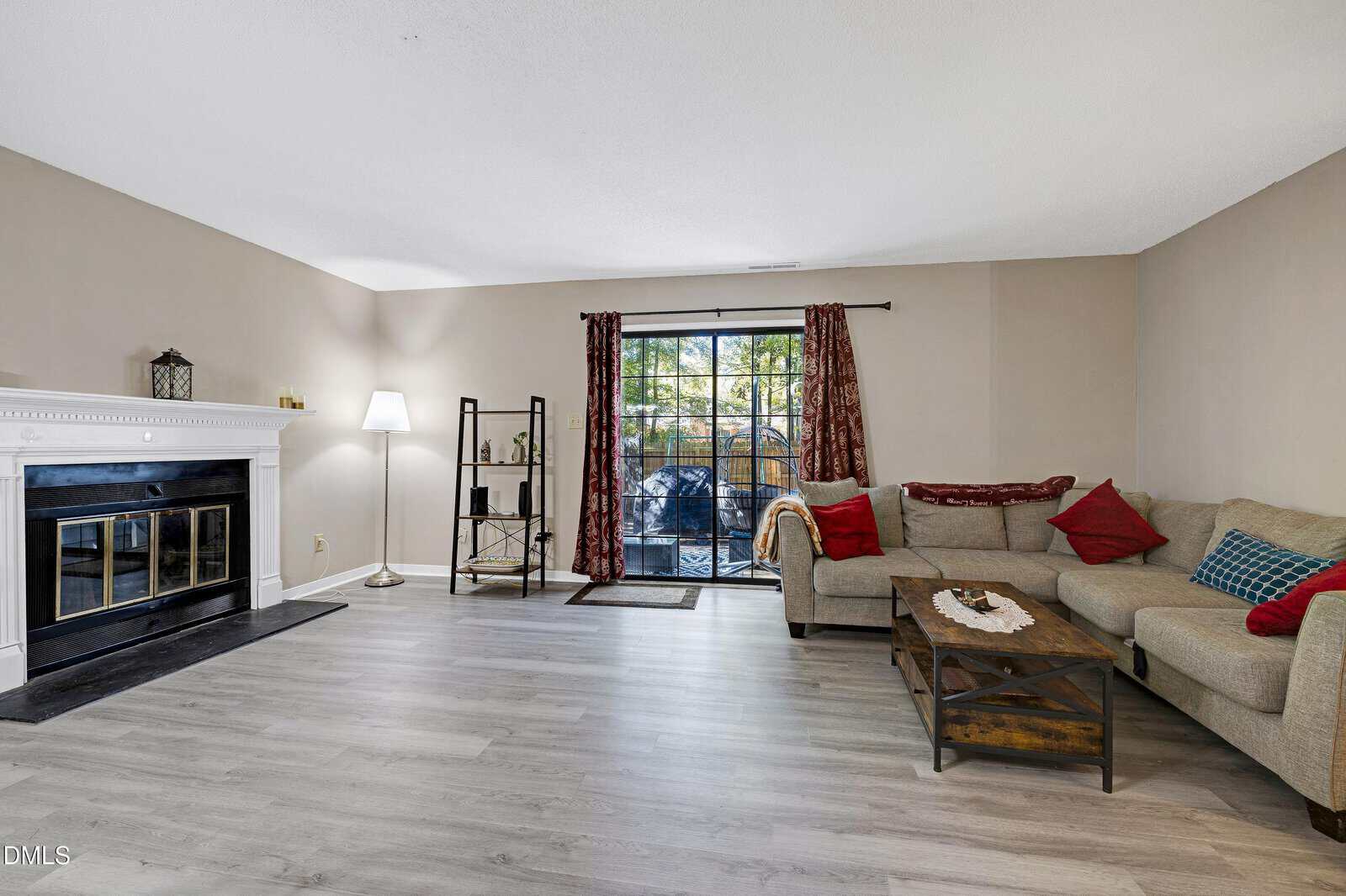 A living room with light wood flooring, a corner sectional sofa with red and blue pillows, a wooden coffee table, a fireplace with a white mantel, a floor lamp, and sliding glass doors with red curtains leading outside.