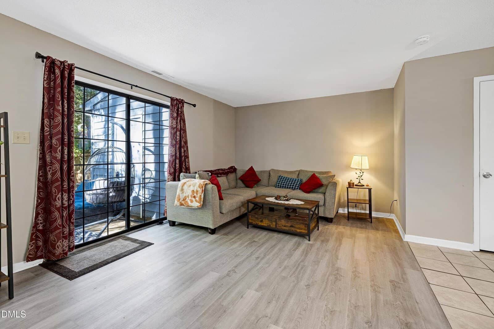 A cozy living room with a sectional sofa, red pillows, a wooden coffee table, a floor lamp, and sliding glass doors leading to a patio with plants. Light wood flooring and neutral walls create a warm atmosphere.
