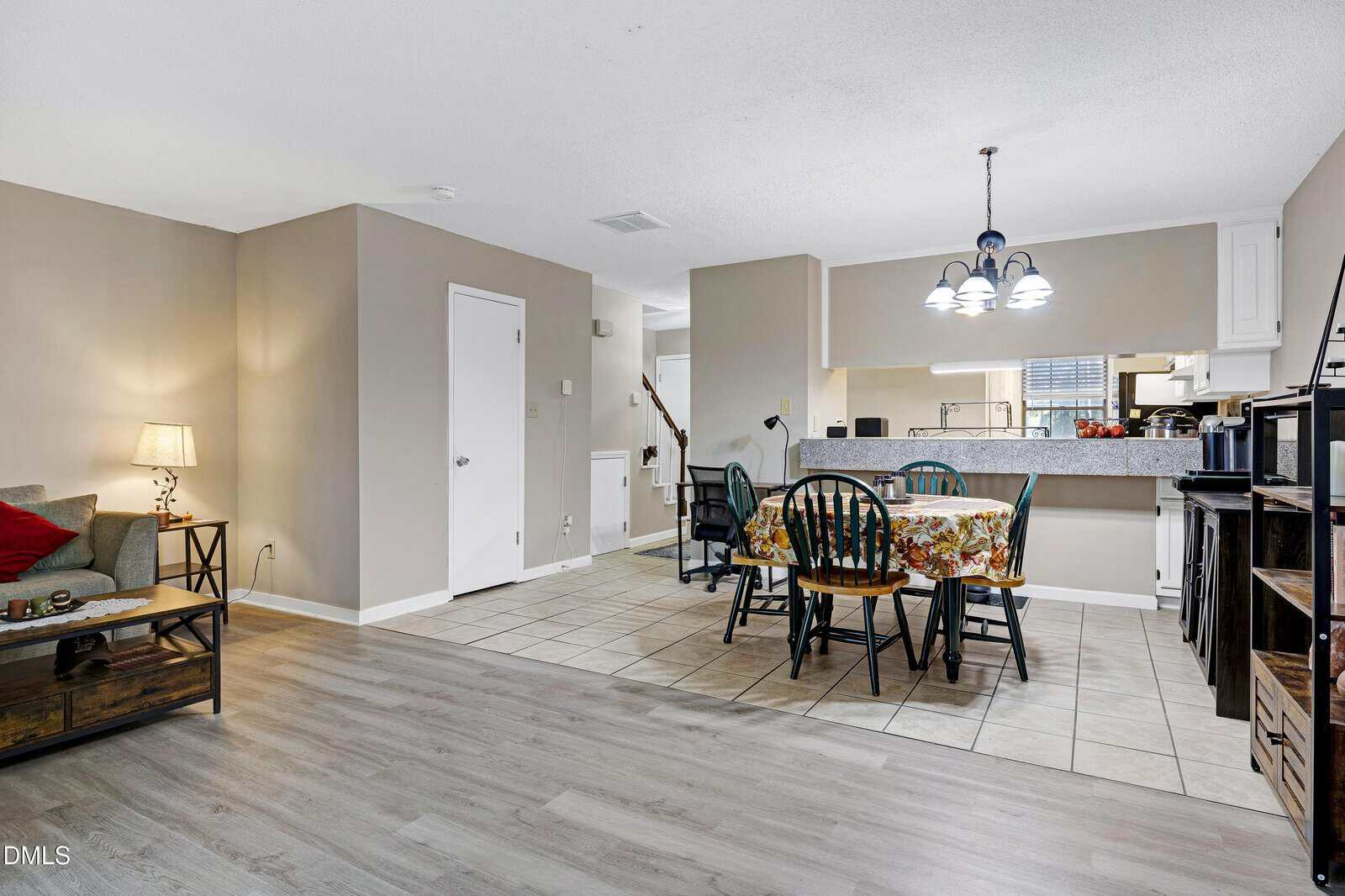 Open dining area with a floral tablecloth, four chairs, tile floor, and kitchen in the background; wood flooring leads to a cozy seating area with a lamp on the left.