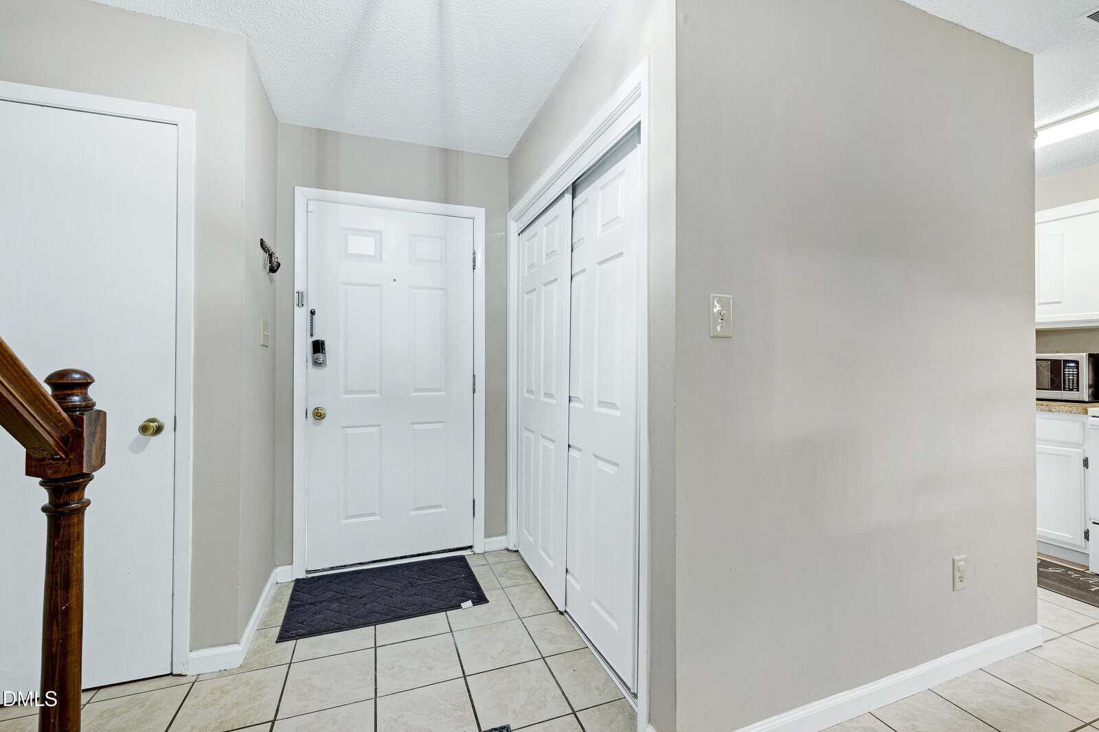 The image shows a tiled entryway with a white front door, two white closet doors, a small black doormat, beige walls, and a partial view of a staircase and kitchen area. Light switch and hook are visible on the walls.