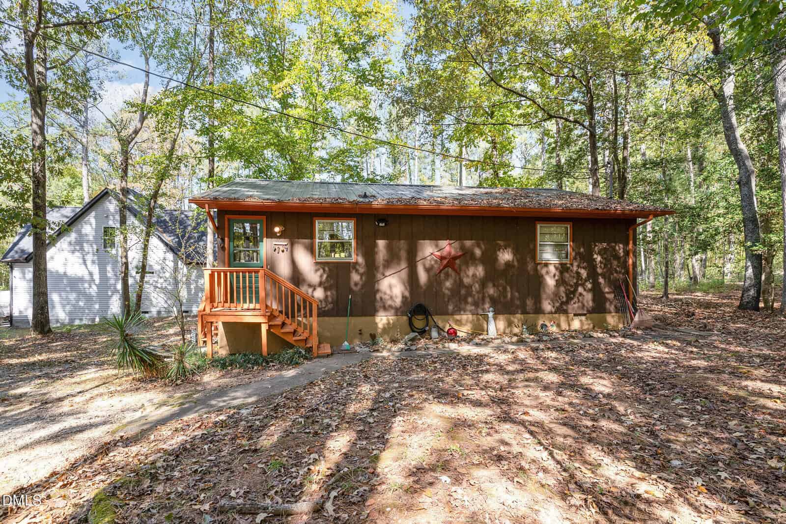 A small, single-story brown house with a covered front porch and two front windows sits in a wooded area at 112 Mohawk Drive in Louisburg. Fallen leaves cover the ground, and another house is partially visible on the left.