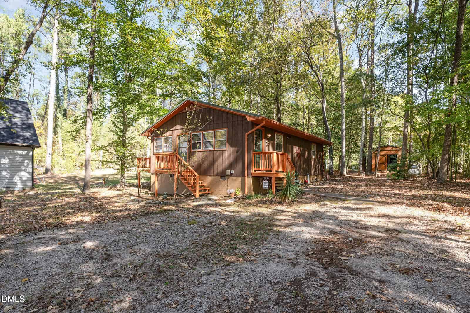 A small brown cabin with a front porch, set on a gravel driveway in a wooded area. Sunlight filters through tall trees surrounding the house. A small outbuilding is visible in the background among the trees.
