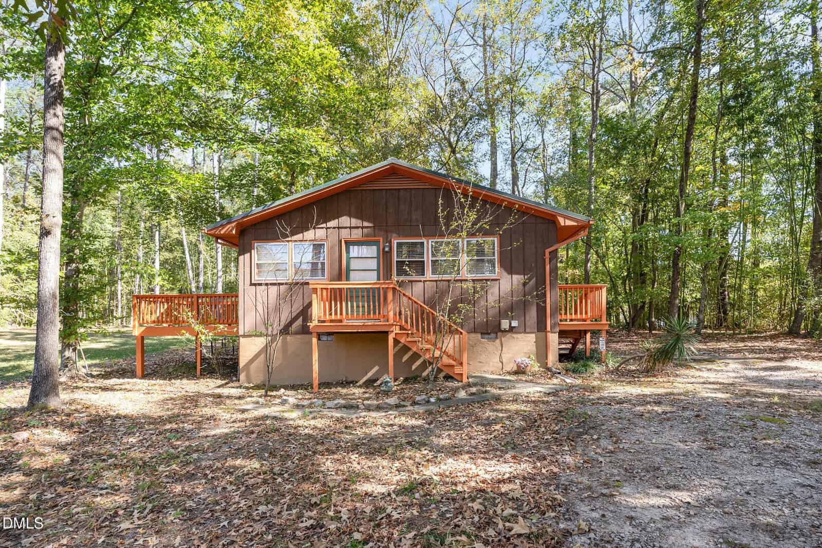 A small brown wooden house with orange trim sits in a wooded area. The house has a front porch, stairs, and large windows. Fallen leaves cover the ground and trees surround the property under a clear sky.