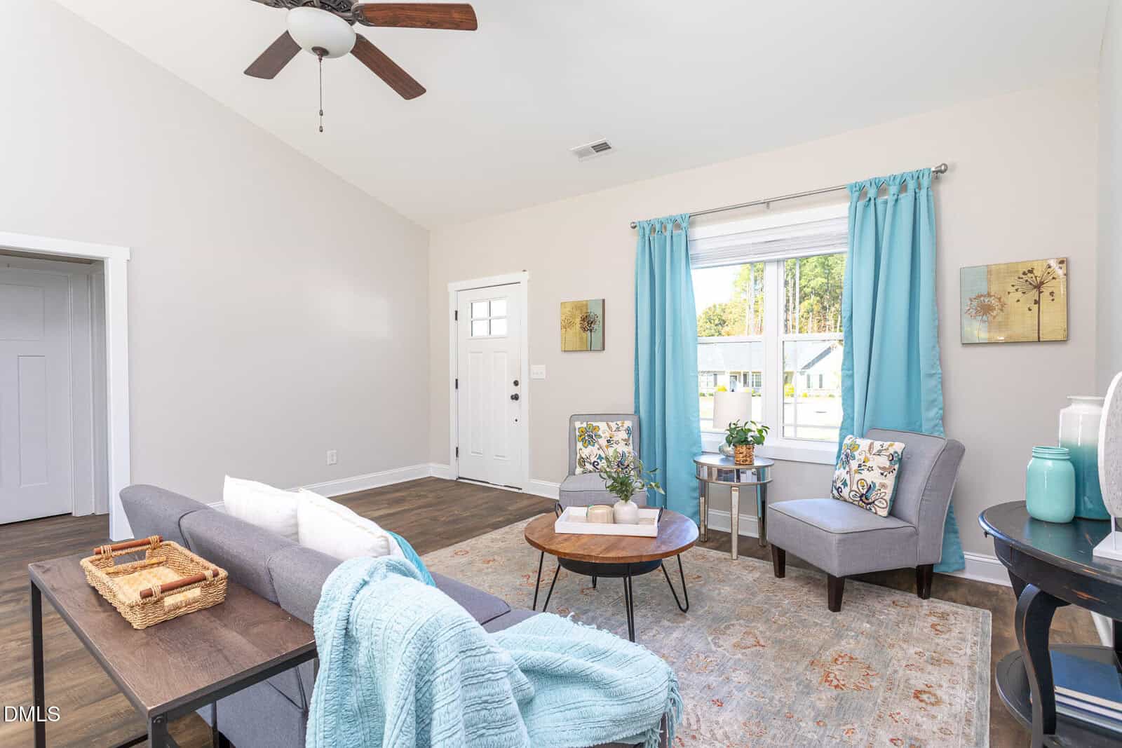 Bright living room with a gray sofa, two gray armchairs, a round coffee table, blue curtains, and a window showing houses outside. Decor includes plants, pillows, and a ceiling fan overhead.