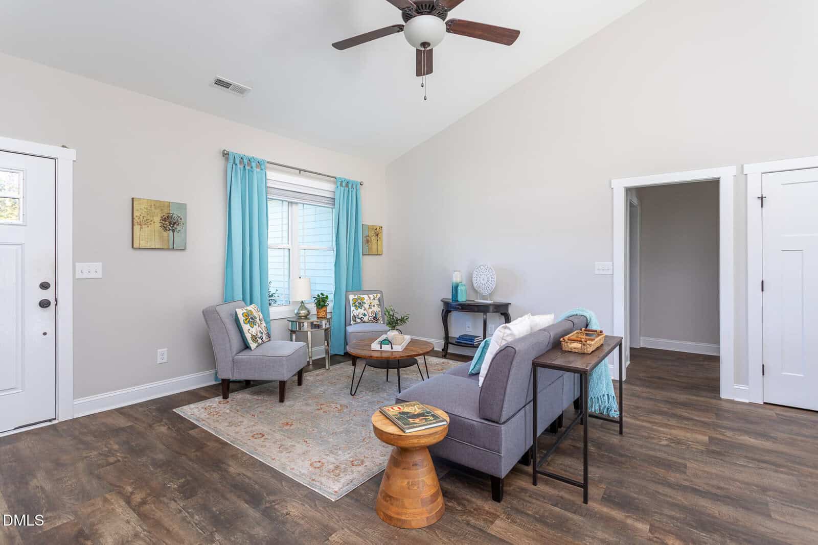 Bright living room with gray furniture, blue curtains, light area rug, wooden floors, ceiling fan, and simple decor. Large window lets in natural light, and a door and doorway are visible in the walls.