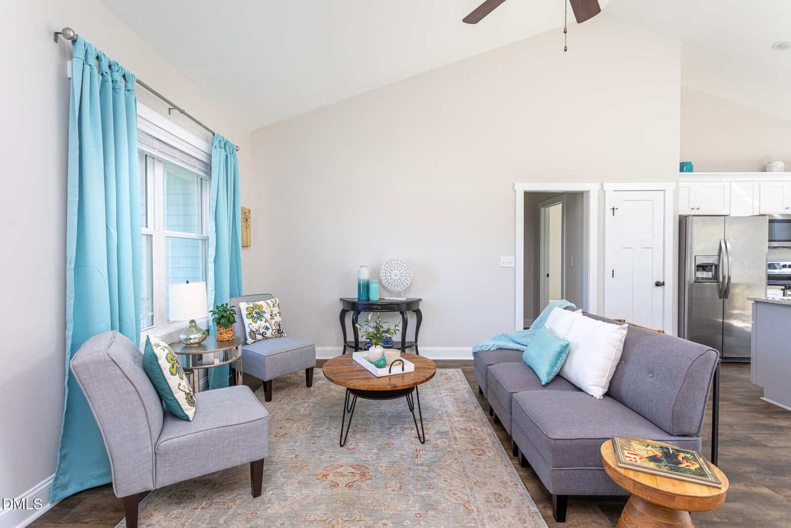 A bright living room with gray armchairs, a gray sofa, a round wooden coffee table, blue curtains, and a neutral area rug. There is a side table with books and a small table against the back wall. The kitchen is visible to the right.