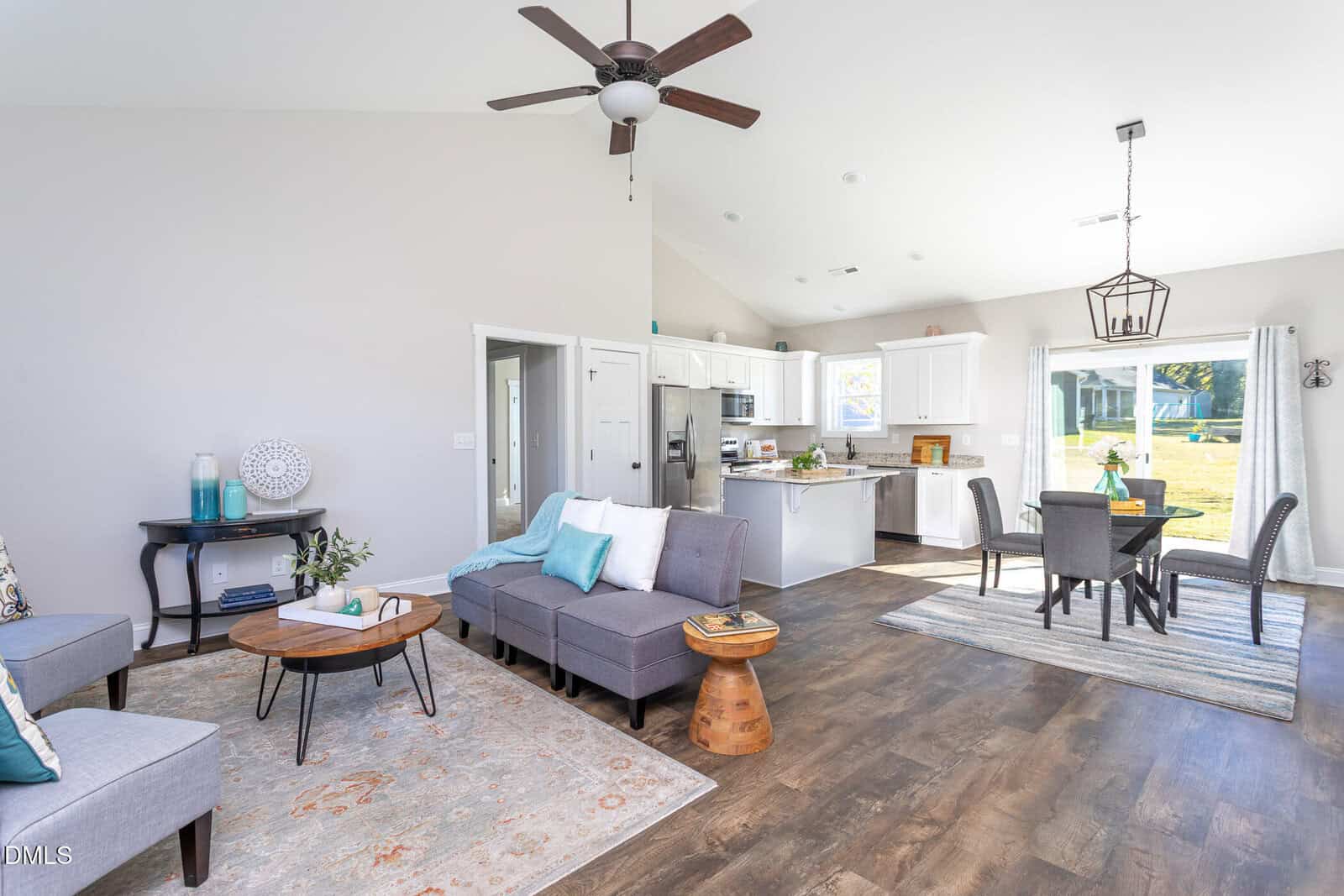 Bright open-concept living and dining area with modern furniture, gray sectional sofa, white kitchen, wooden floors, large windows, and a ceiling fan. Sliding doors open to a sunny backyard.