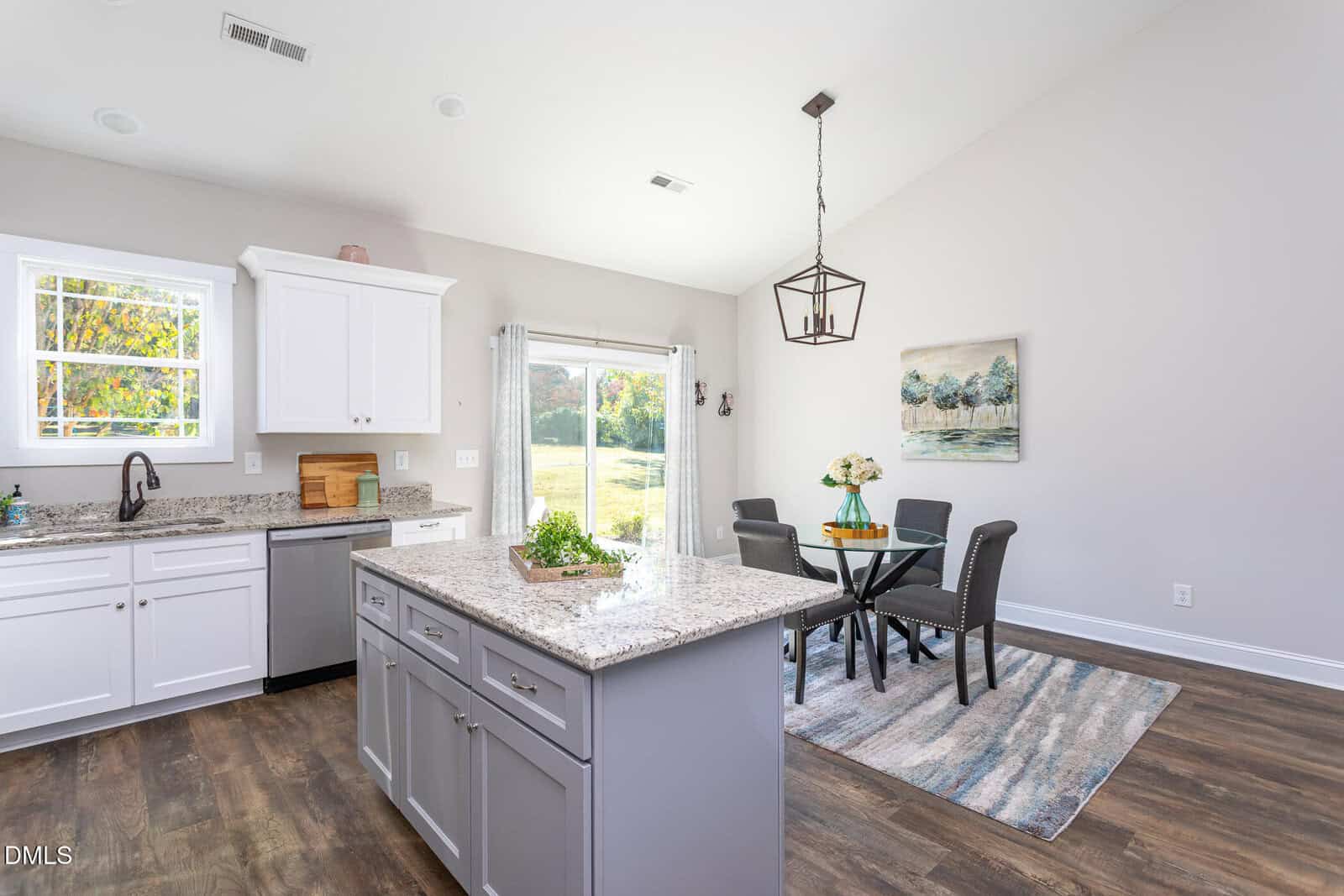 Bright, modern kitchen with white cabinets, a gray island, granite countertops, and stainless steel fixtures. Dining area with a round table and four chairs sits on a rug near large sliding doors to a sunny backyard.