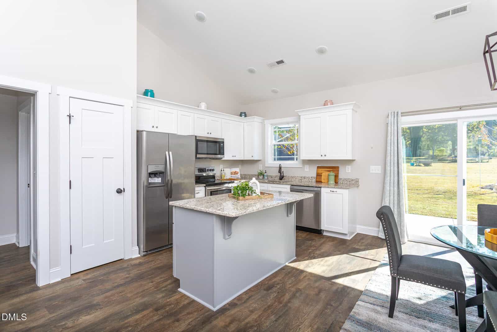 Bright, modern kitchen with white cabinets, stainless steel appliances, granite countertops, and a central island. Sliding glass doors open to a sunny backyard, and a dining area with gray chairs sits nearby.