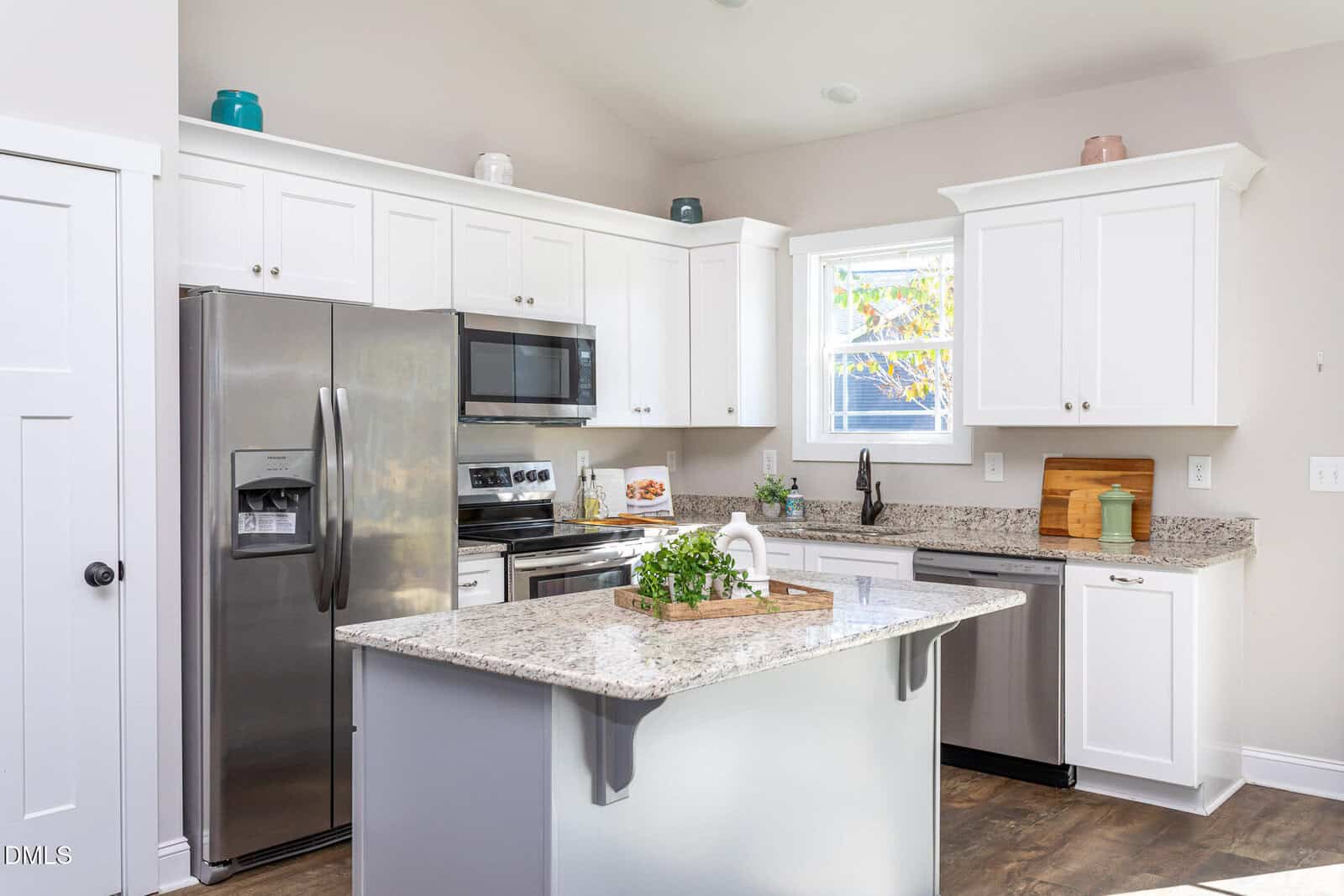 Modern kitchen with white cabinets, stainless steel appliances, a granite island with a small plant, and decorative jars on top of cabinets. Sunlight streams through a window above the sink.