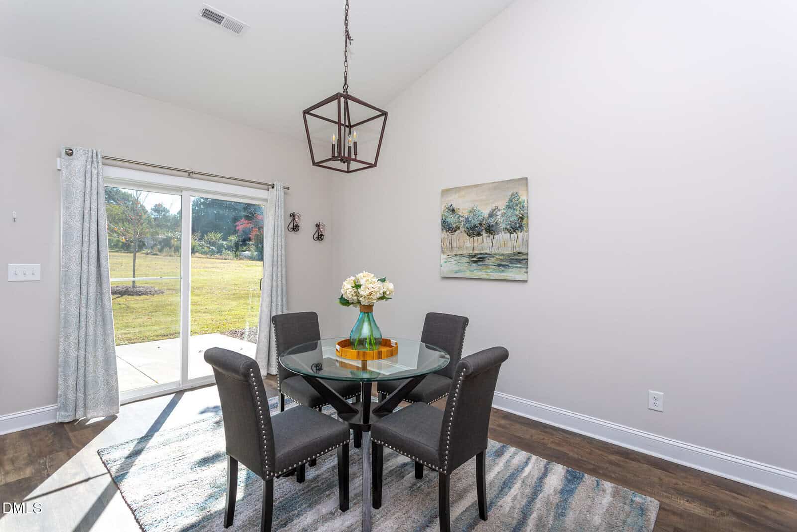 A modern dining area with a round glass table, four upholstered chairs, a hanging light fixture, sliding glass doors with curtains, a rug, wall art, and a vase with flowers, overlooking a grassy backyard.