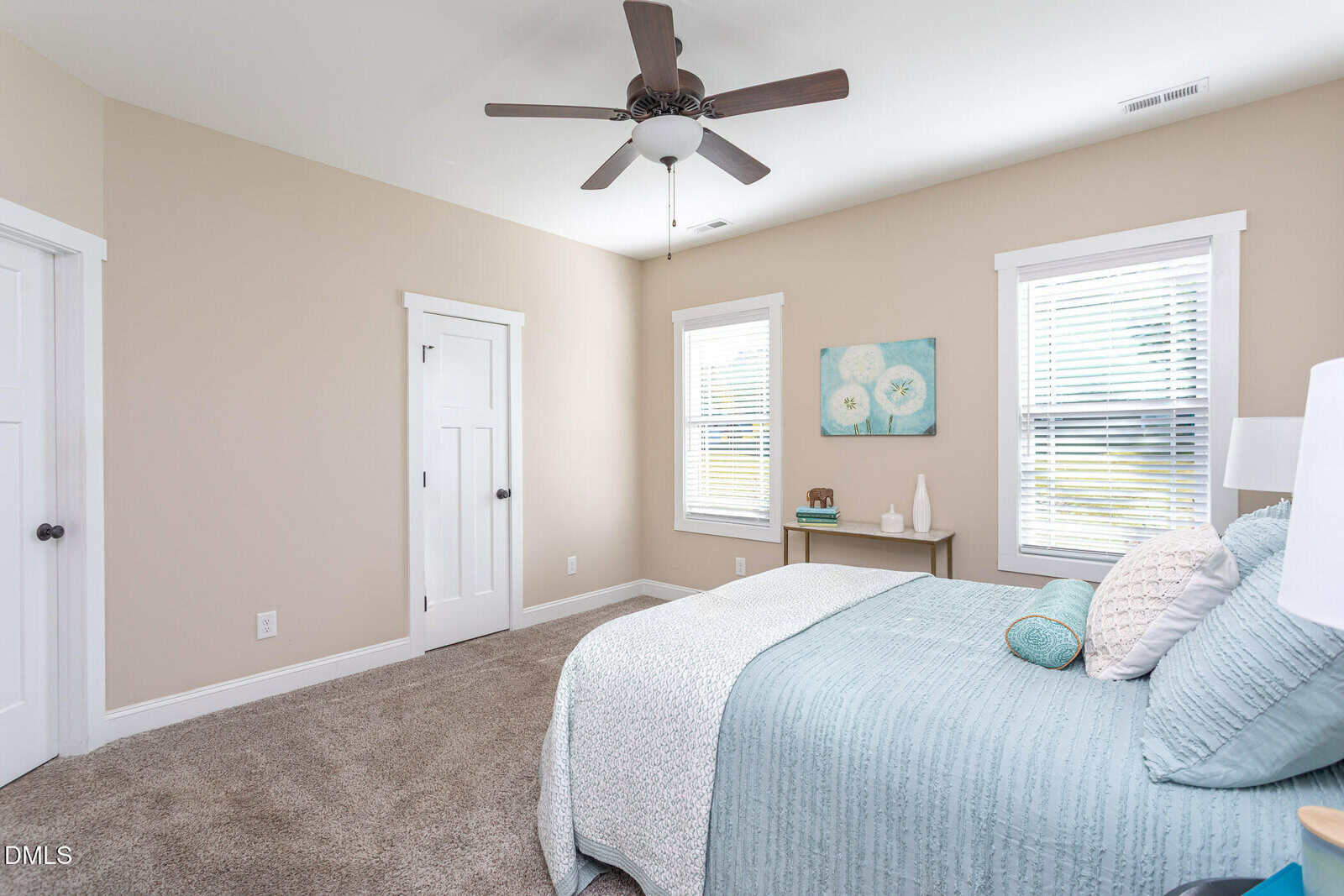 A bedroom with beige walls, two windows, a bed with blue bedding, a ceiling fan, a nightstand with decor, and closed white doors. A floral painting hangs above the bed. Natural light fills the room.