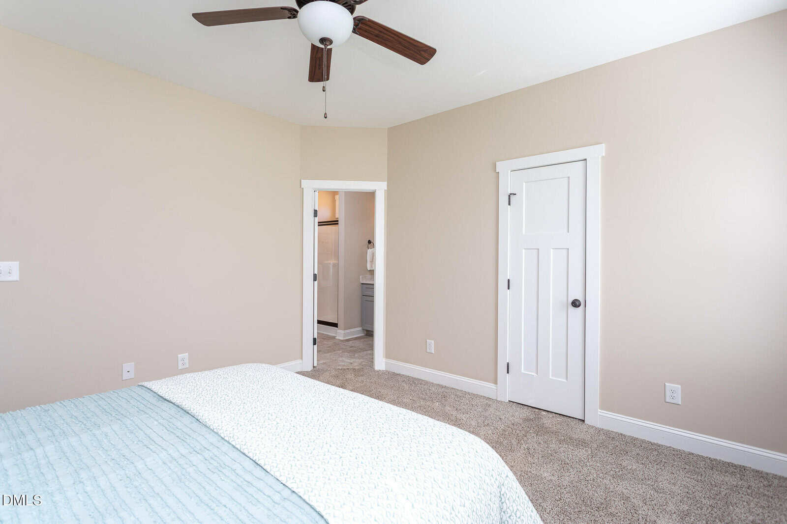 A bedroom with beige walls, carpeted floor, a bed partially visible, ceiling fan, a white door, and an open doorway leading to a bathroom with a sink and mirror visible.