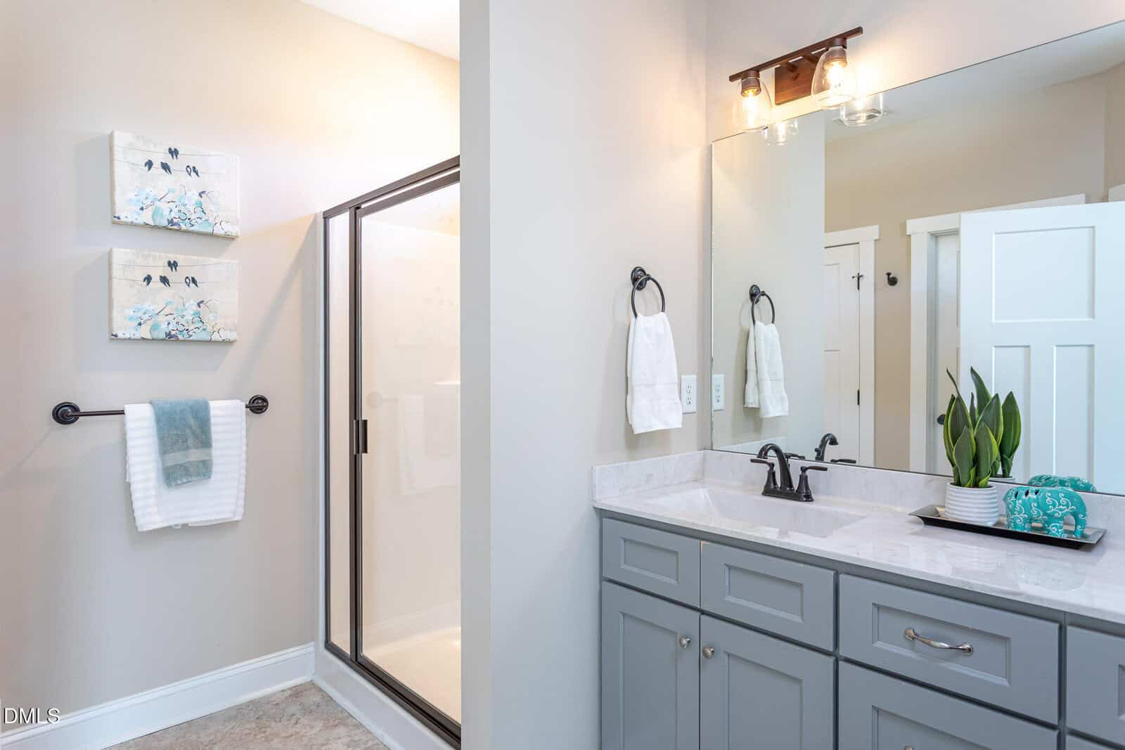 Bright bathroom with a glass shower, gray vanity with black hardware, white countertop, towels hanging on racks, decorative paintings, and a potted plant near the sink.