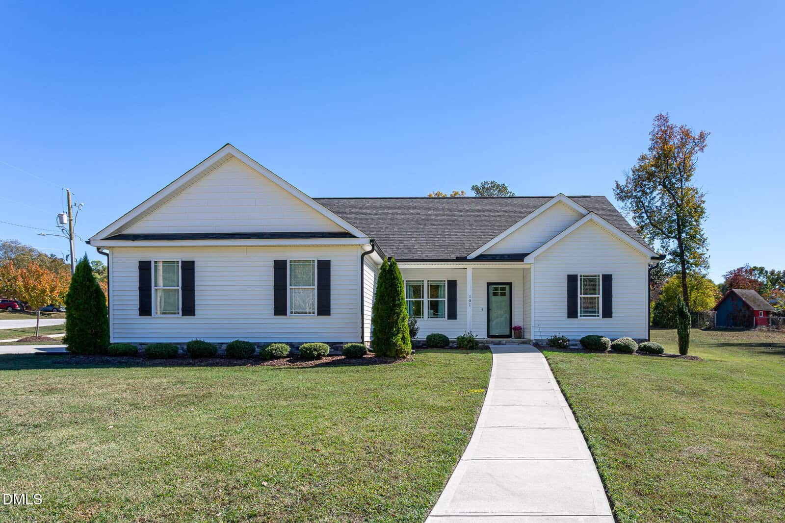 A single-story white house with black shutters, a dark gray roof, and a light green front door sits at 101 Briarwood Court in Louisburg. A concrete walkway leads to the entrance, surrounded by lawn, shrubs, and trees under a clear blue sky.