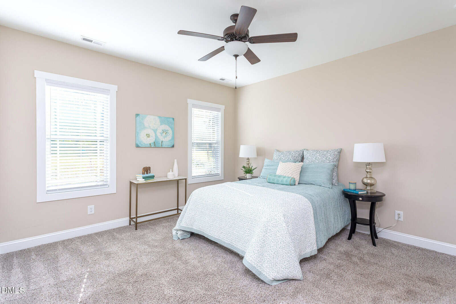 A bright bedroom with beige walls, a bed with light blue and white bedding, two white-shaded lamps on side tables, art on the wall, a ceiling fan, and two windows with white blinds letting in natural light.