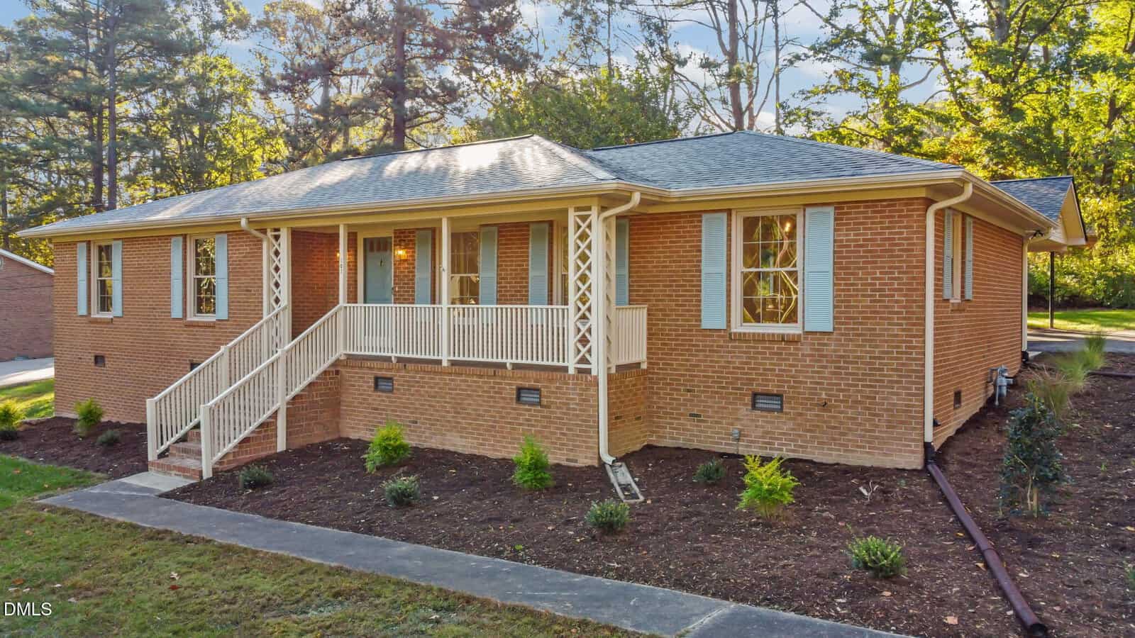 A single-story brick house at 102 Royall Road, Oxford, features light blue shutters, a covered front porch with white railings and stairs. The landscaped yard has mulch, small plants, and tall trees in the background.