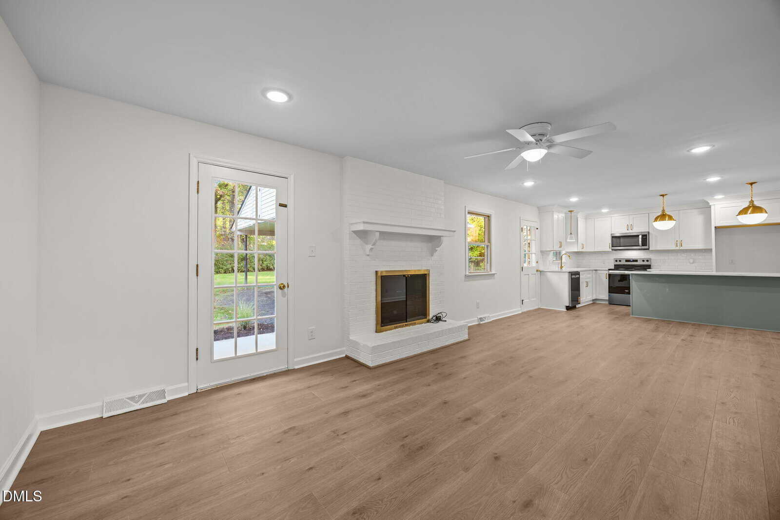 Open-plan living area with light wood flooring, white walls, a brick fireplace, ceiling fan, recessed lighting, and a door leading outside. The kitchen features white cabinets, stainless steel appliances, and pendant lights.