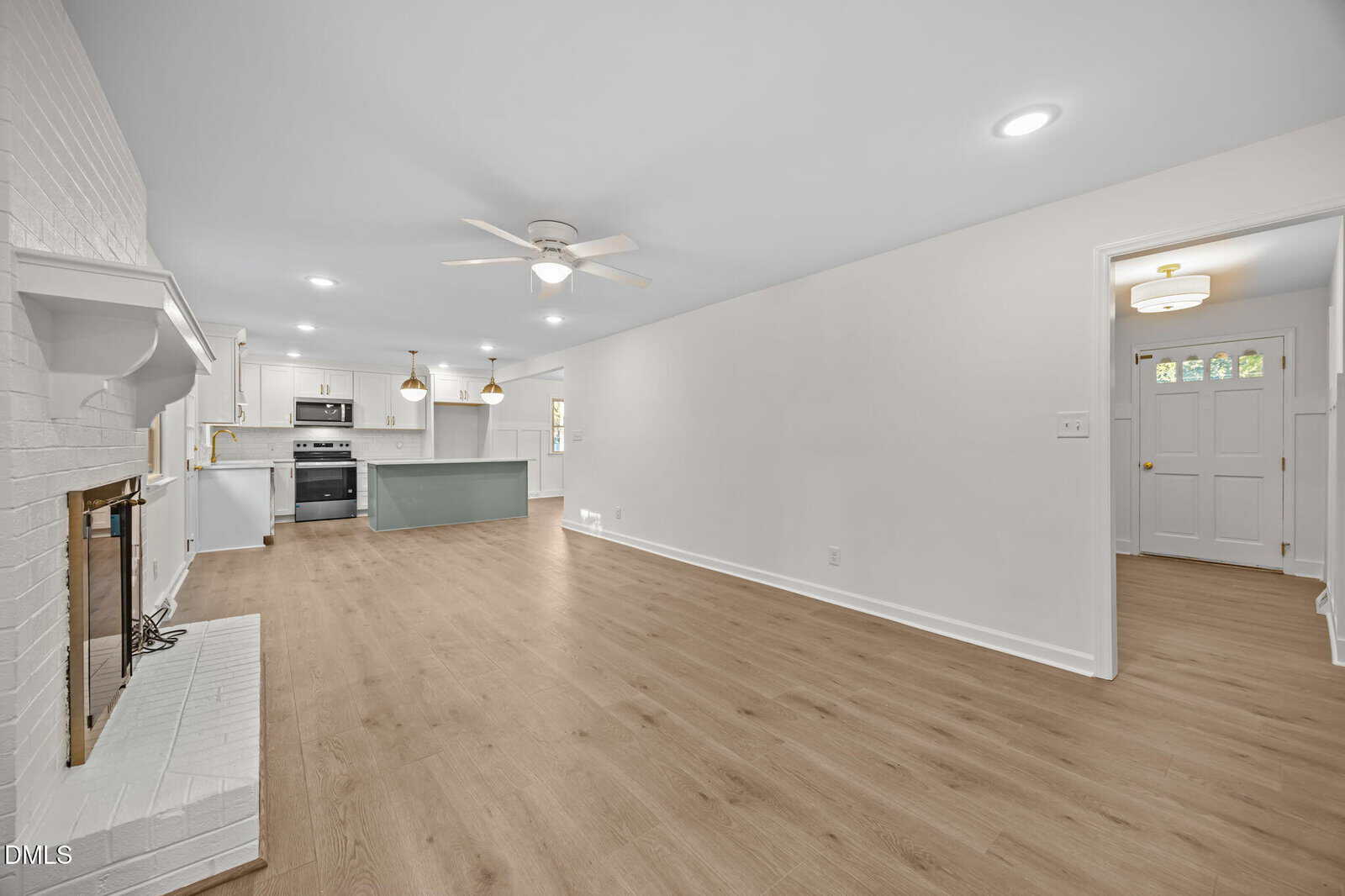 Open-concept living room with light wood flooring, white walls, ceiling fans, recessed lighting, a white brick fireplace, and a view into a modern kitchen with an island and stainless steel appliances. Entryway visible on the right.
