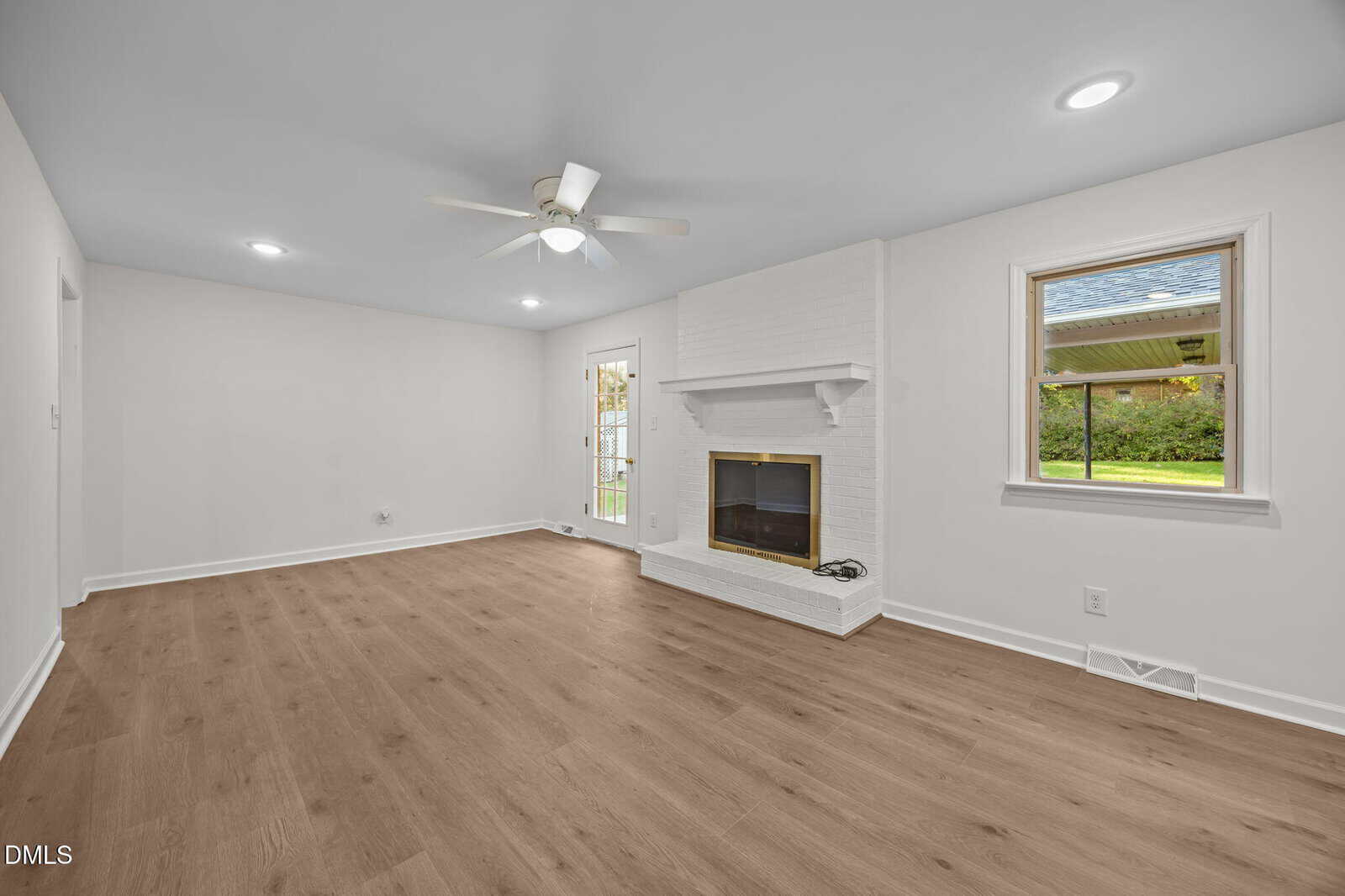 A bright, empty living room with light-colored walls, wood-style flooring, a white ceiling fan, recessed lighting, a white brick fireplace, a small window, and a glass door opening to the outside.