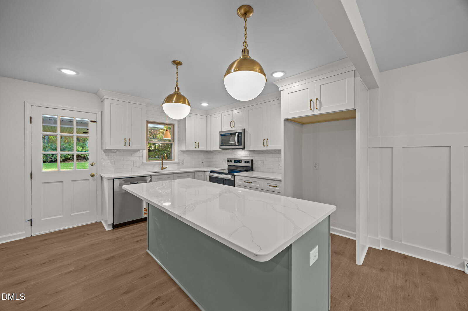 A modern kitchen with white cabinets, stainless steel appliances, a center island with a gray base and white countertop, wood flooring, brass light fixtures, and a door leading outside.