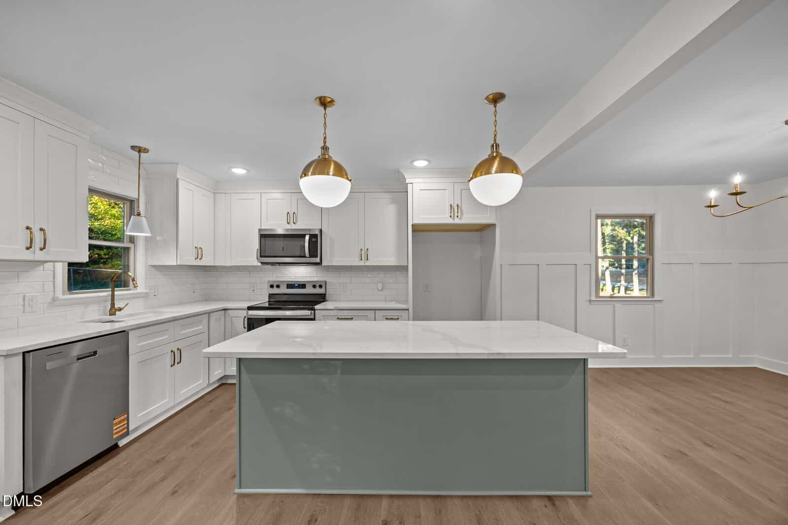 Modern kitchen with white cabinetry, stainless steel appliances, light wood flooring, a large island with a gray base and white countertop, two gold pendant lights, and windows letting in natural light.