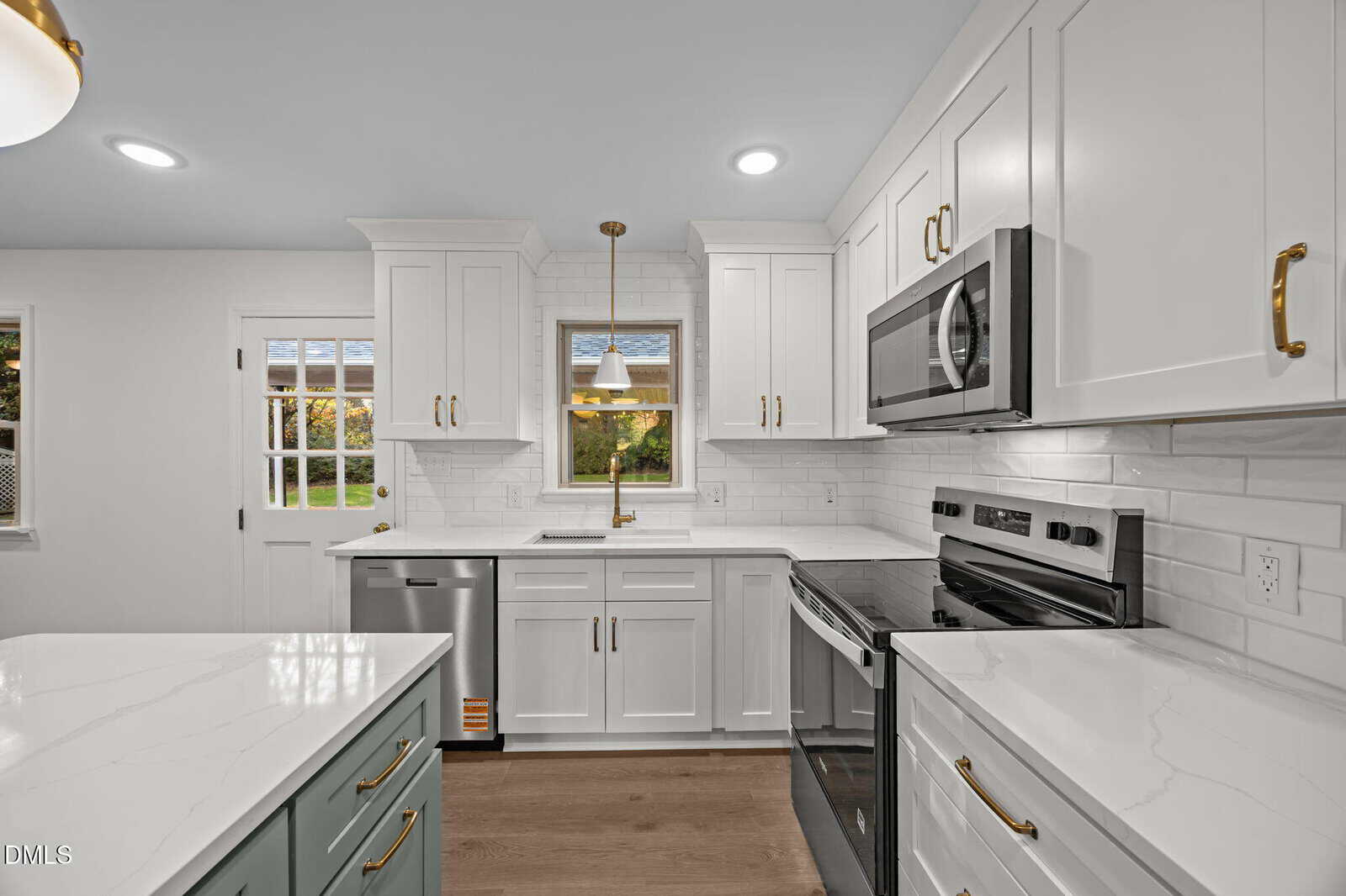 Modern kitchen with white cabinets, stainless steel appliances, gold hardware, a farmhouse sink under a window, white subway tile backsplash, and marble countertops. Light wood flooring and a small island are visible.