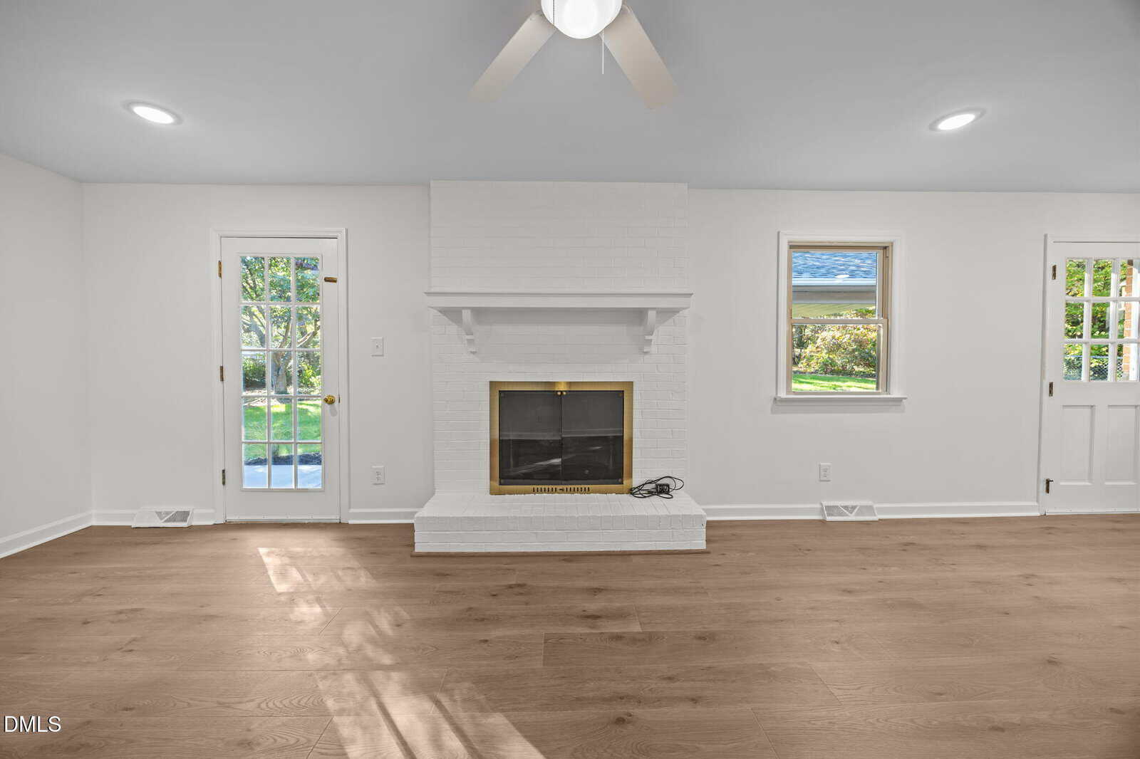 A bright living room with light wood floors, a white brick fireplace with a brass-trimmed glass door, a ceiling fan, and large windows and doors allowing natural light to enter.