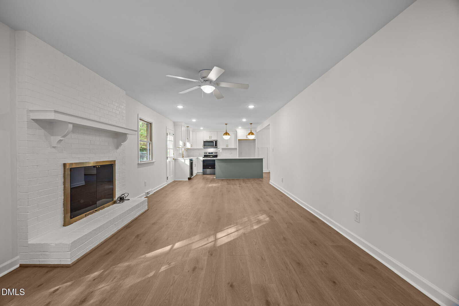 A spacious open-concept living area with light wood floors, white walls, a white brick fireplace on the left, recessed lighting, ceiling fan, and a modern kitchen with island and pendant lights in the background.