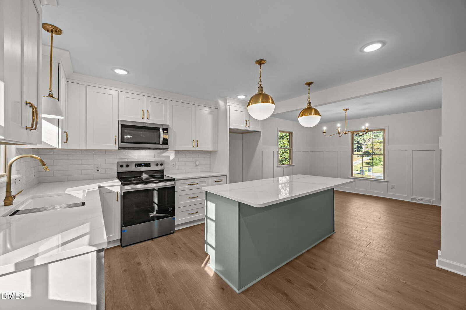 Modern kitchen with white cabinets, stainless steel appliances, a green island with a white countertop, gold hardware, pendant lights, hardwood floors, and large windows letting in natural light.