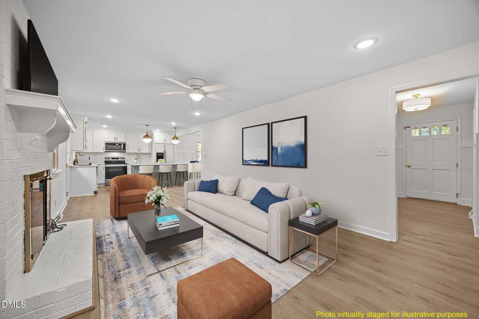 A bright, modern living room with a white brick fireplace, beige sofa, abstract wall art, coffee tables, and a rug. The open layout leads to a white kitchen with bar stools. Text in the bottom right notes virtual staging.