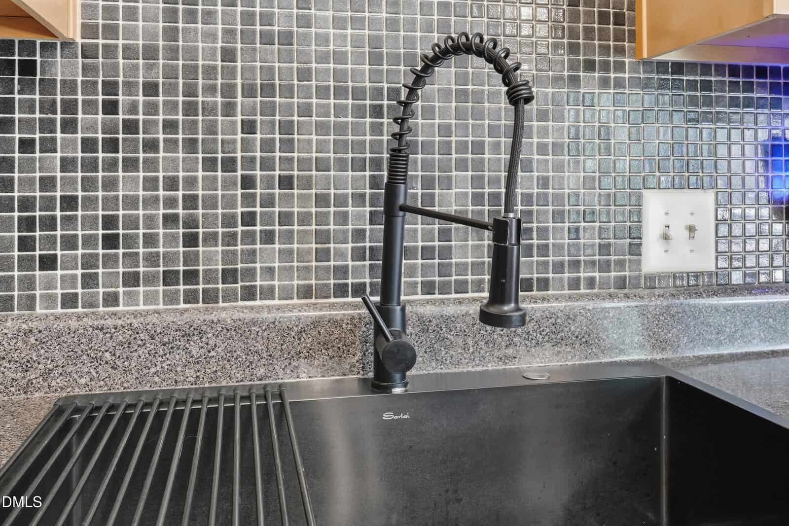A modern black kitchen sink with a spring coil faucet is set against a backsplash of small black and gray tiles. The countertop is dark and speckled, with an integrated draining area on one side.