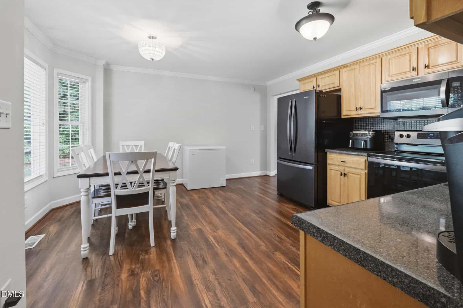 A kitchen with wood flooring, light wood cabinets, black appliances, and a black countertop. A dining table with white chairs sits near a window, and a white chest freezer is against the wall.