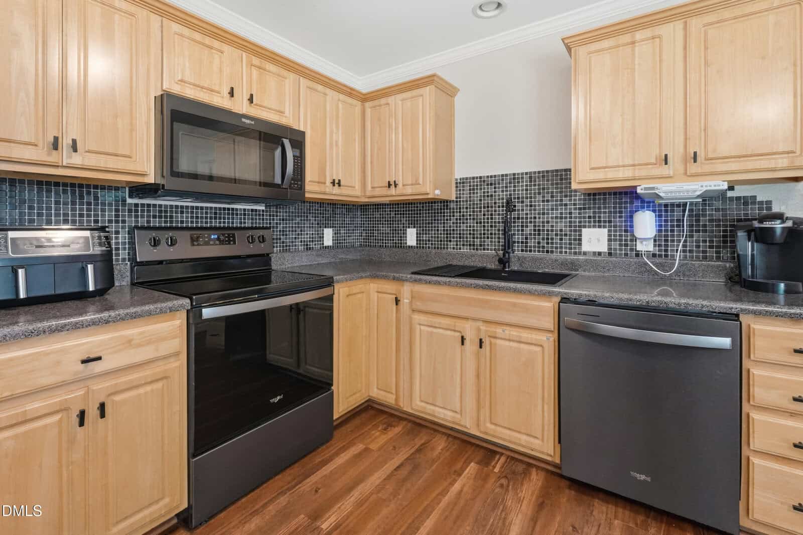 A modern kitchen with light wood cabinets, black appliances including a stove, microwave, and dishwasher, a black tile backsplash, countertop appliances, and a dark sink, all set on a wood floor.